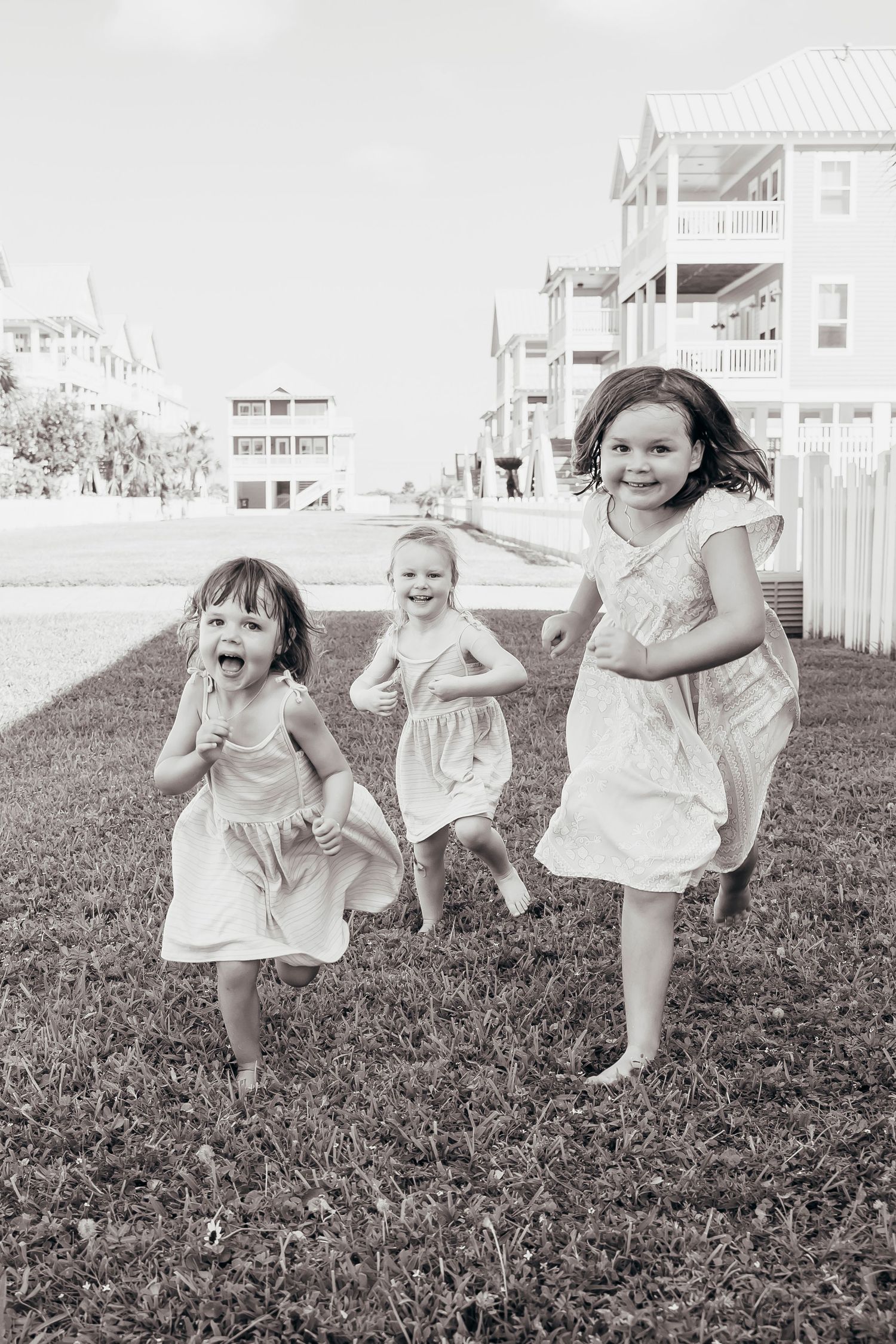 Three young girls in dresses running and laughing on grass near beach houses in black and white photo.
