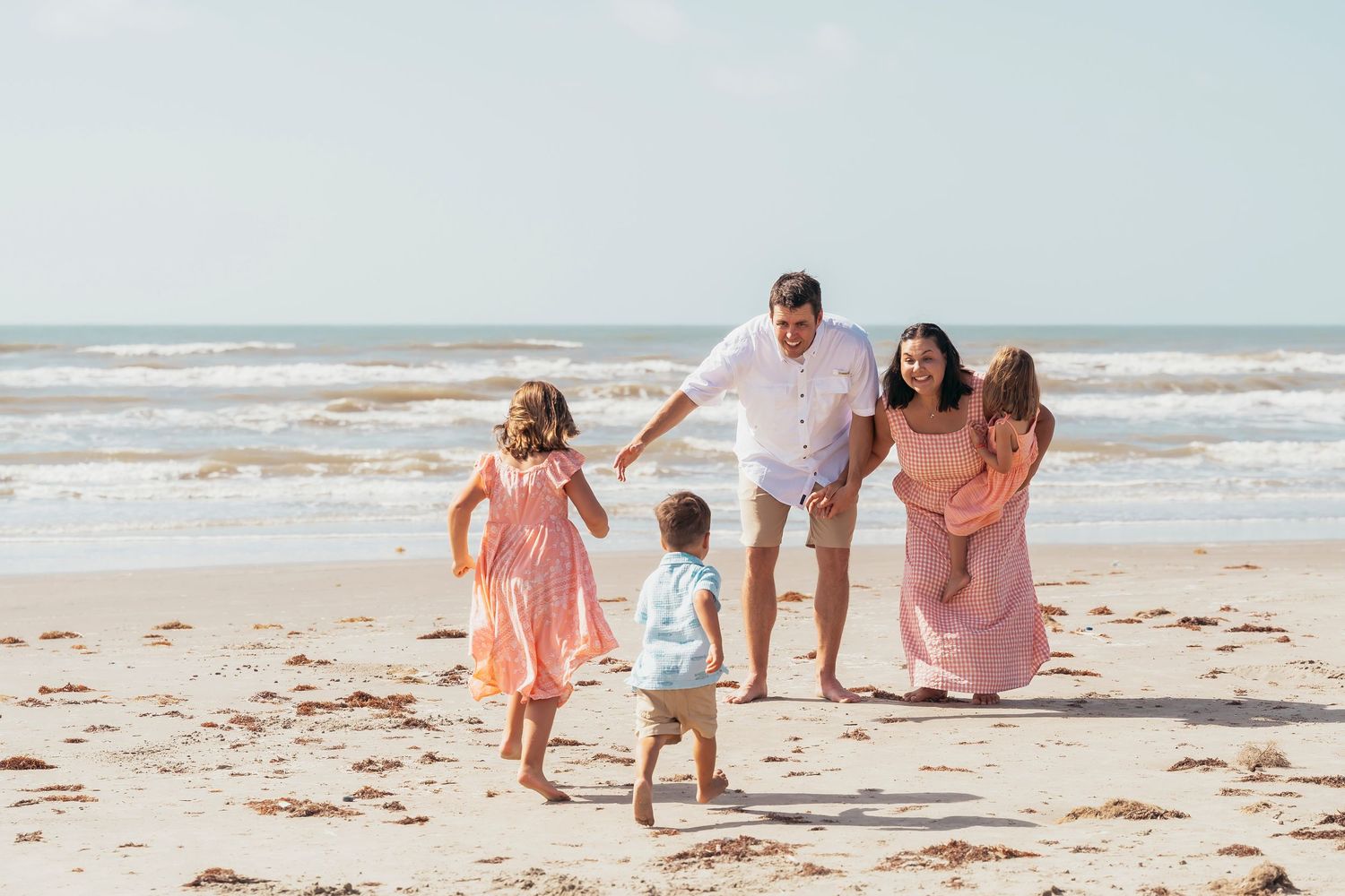 Happy family playing on a sunny beach, parents with three young children running toward them near the ocean.