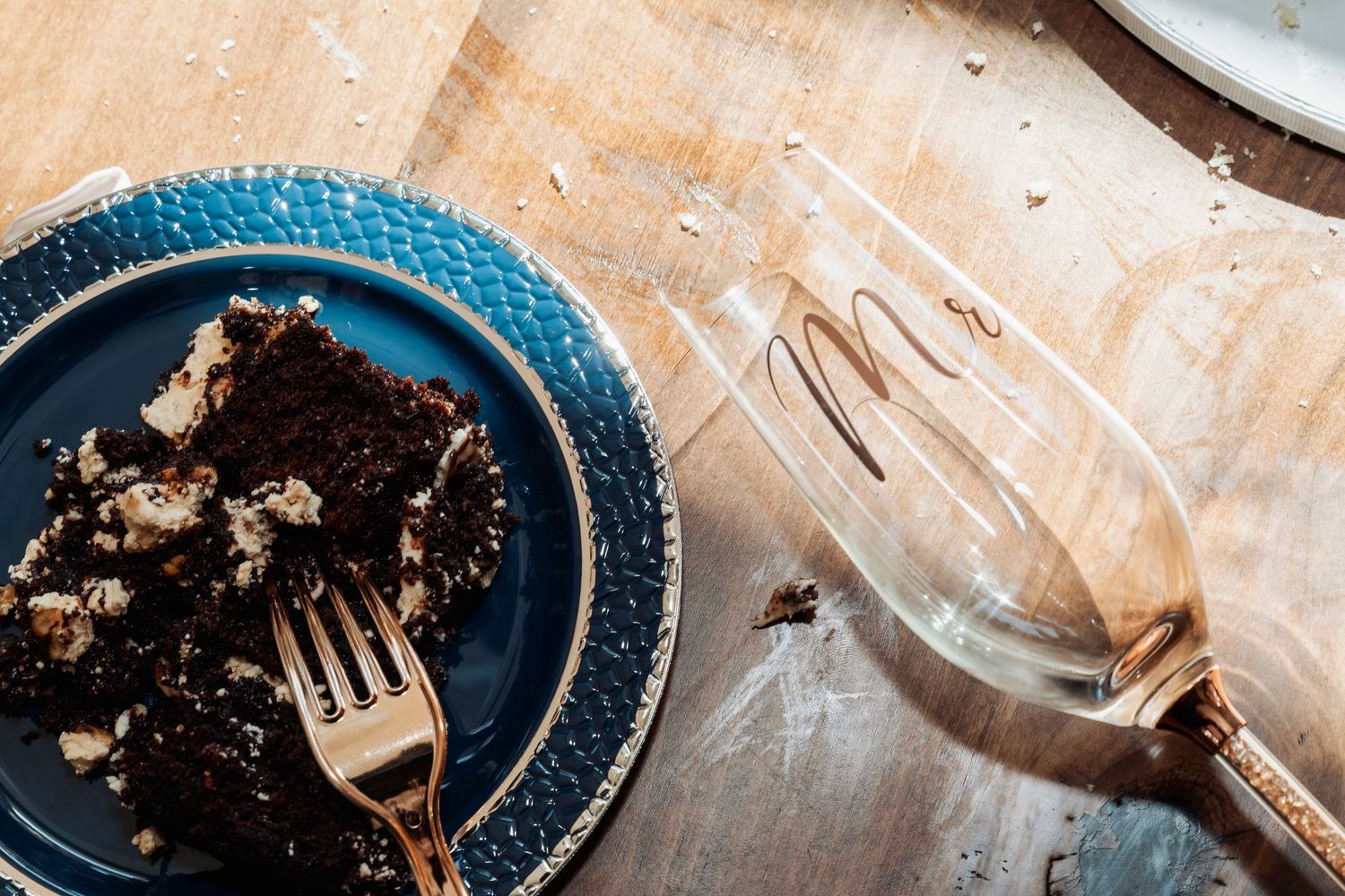 Chocolate cake slice on blue plate with fork beside a decorative glass on wooden table.