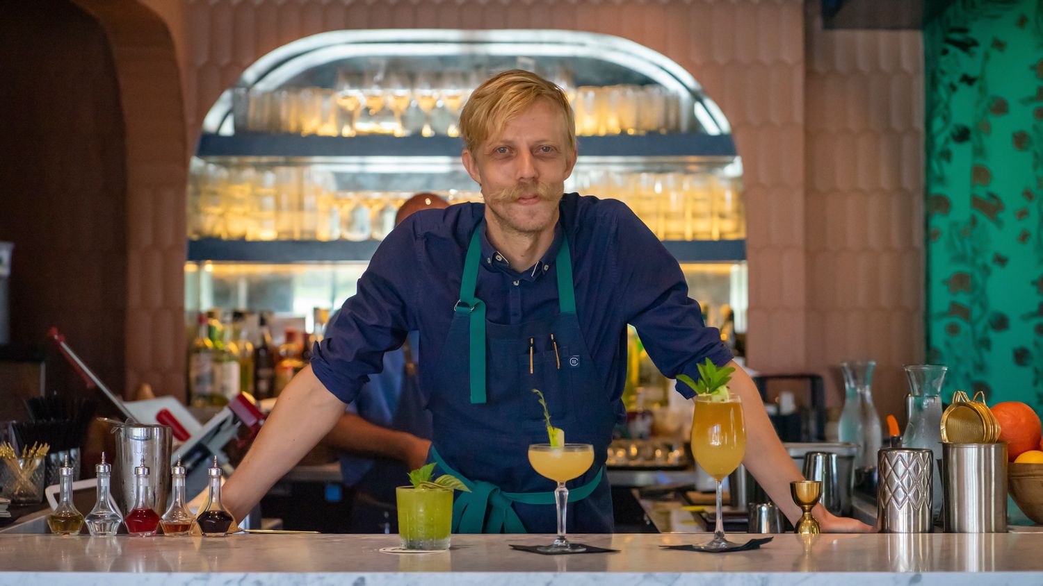A smiling bartender in a teal apron poses behind a bar with freshly crafted cocktails on the counter.