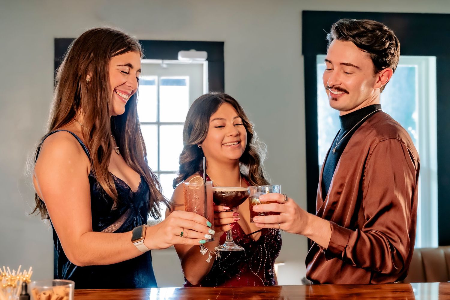 Three friends smiling and toasting with cocktails at a bar in elegant evening attire.