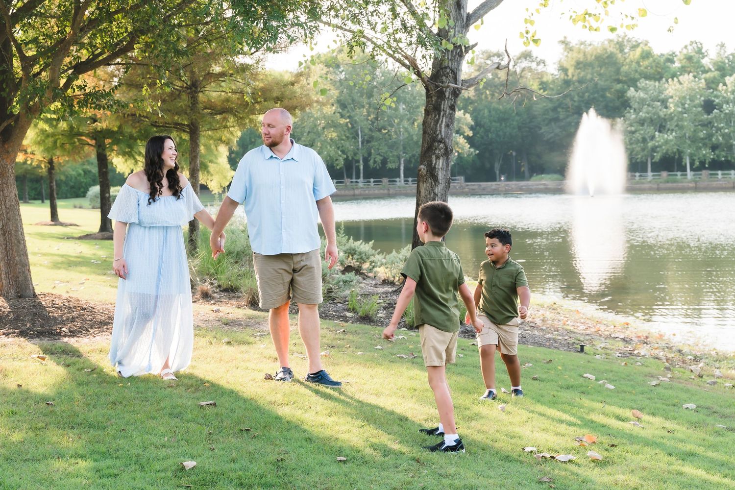 Family of four walking hand-in-hand by a serene pond in a lush green park on a sunny day.