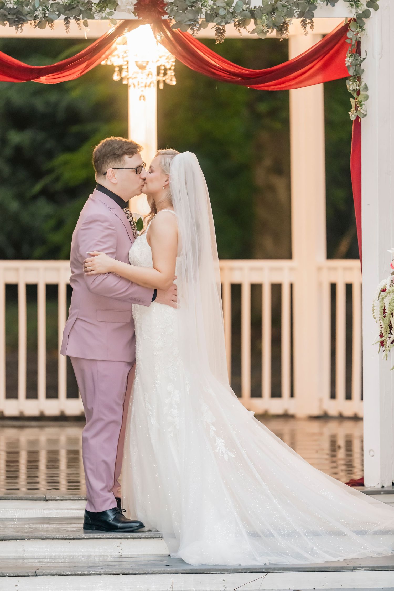 Bride and groom share first kiss under white gazebo with red draping at outdoor wedding ceremony.
