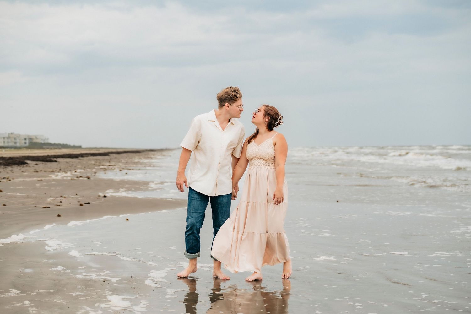 A couple walks barefoot on a beach, waves lapping at their feet, sharing a romantic gaze in soft natural light.