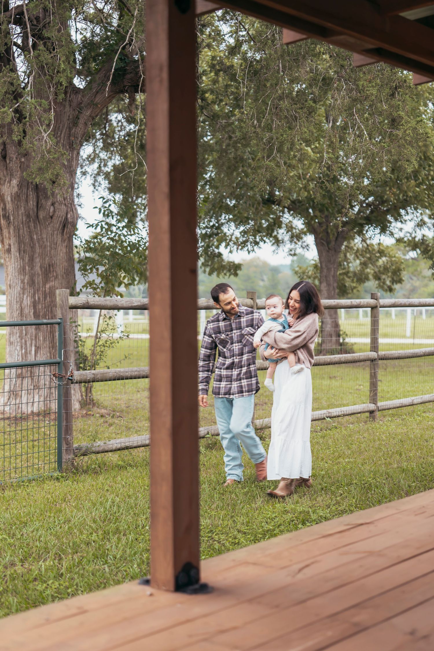 A young family with a baby enjoys a sunny day on a rustic farm, standing near a wooden fence surrounded by trees.