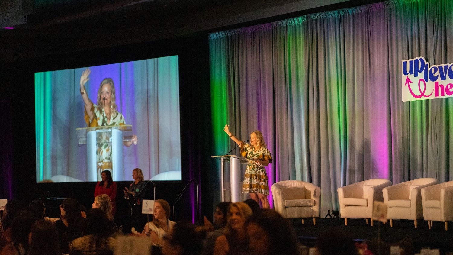 Woman speaker on stage at Uplevel Her conference, gesturing enthusiastically at podium with large screen projection behind her.