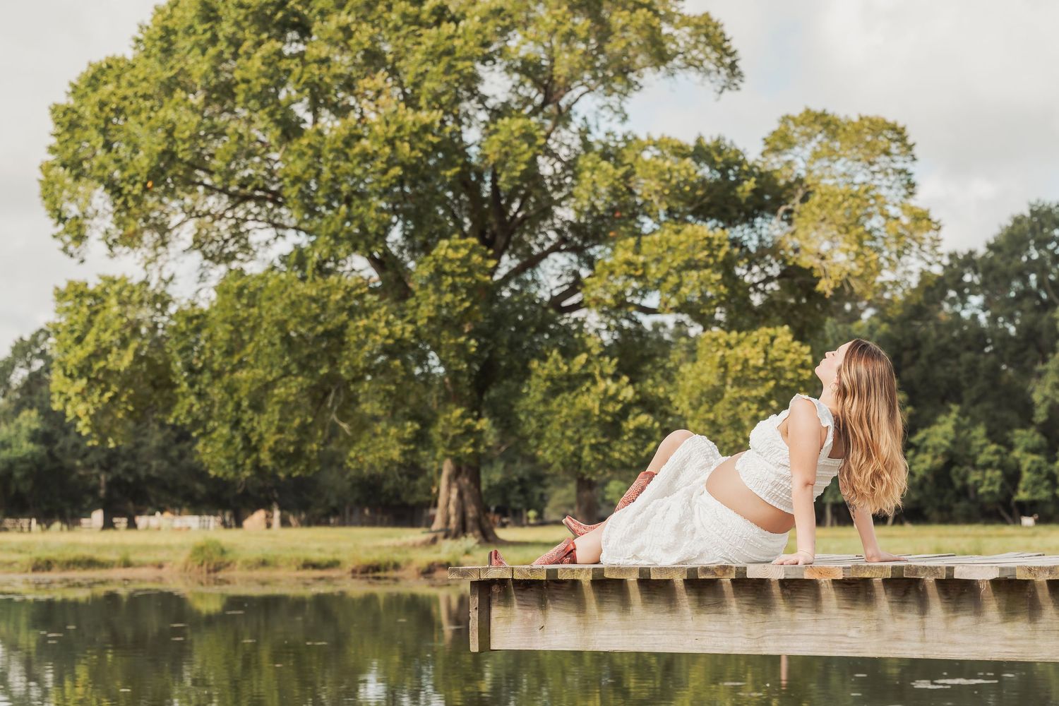 Pregnant woman in white dress relaxing on wooden dock by calm lake surrounded by lush green trees.