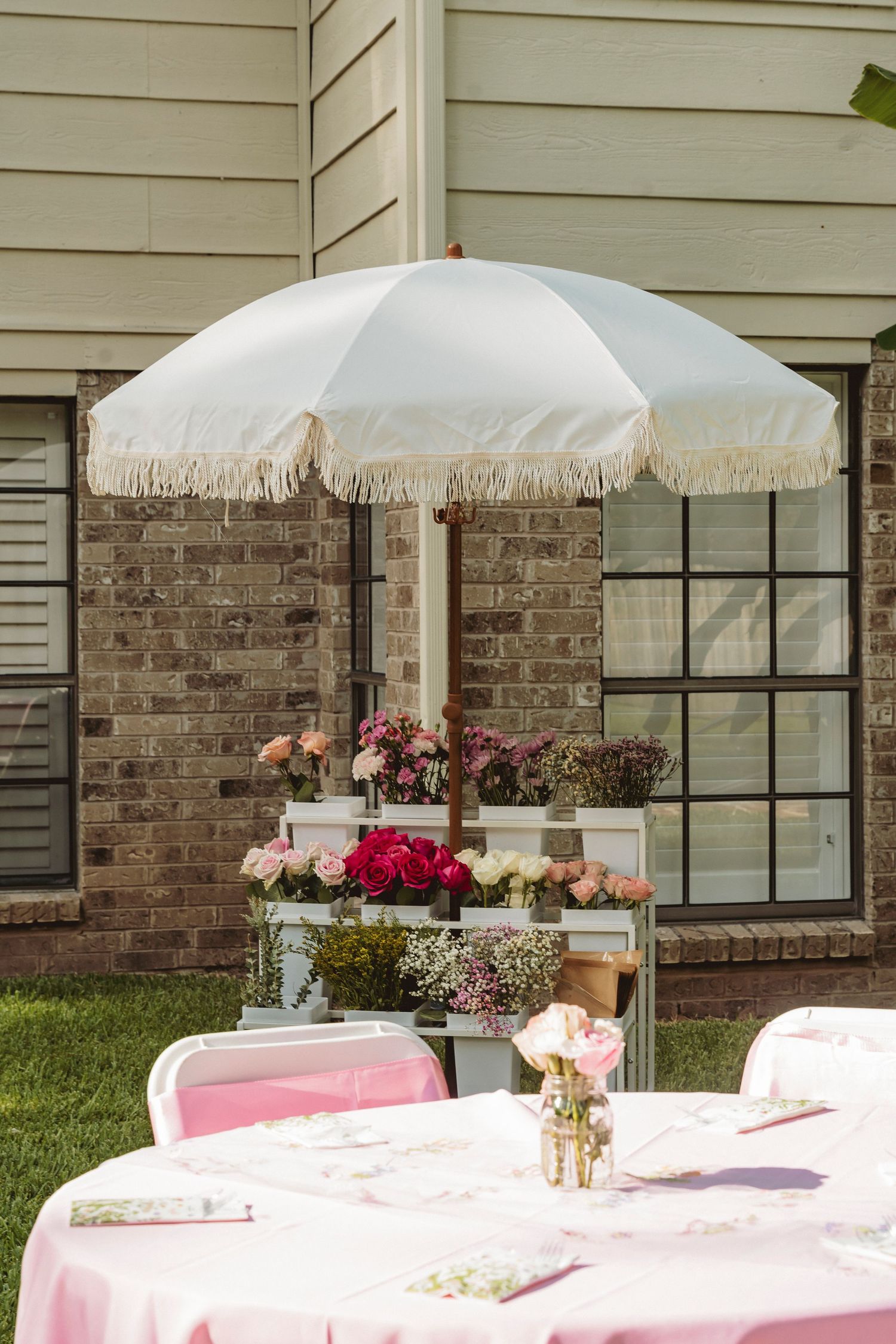 Elegant outdoor garden party setup with white fringed umbrella, pink tablecloth, and floral arrangements near brick building.