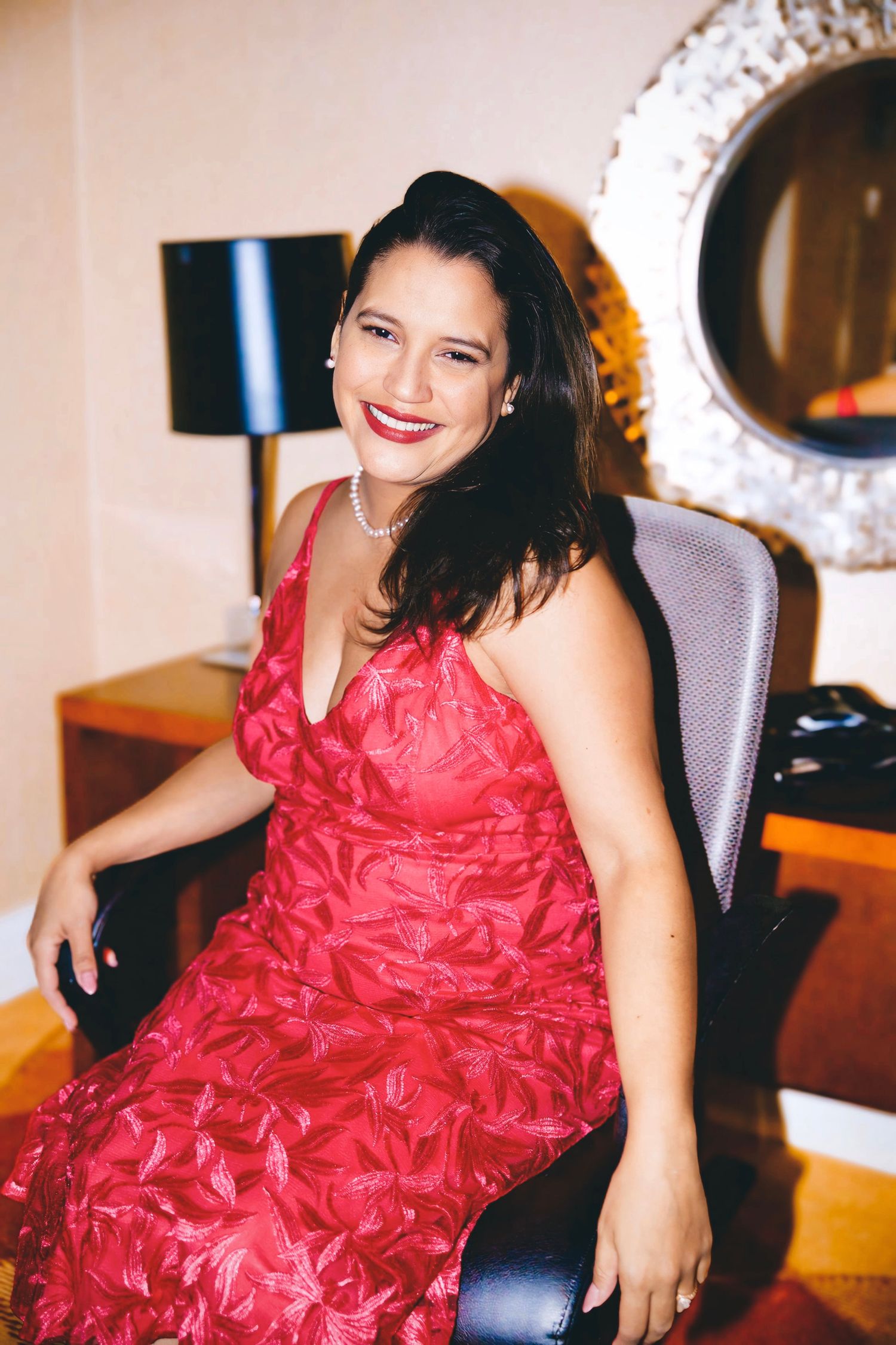 Portrait of a smiling woman in a red dress seated indoors, styled with pearls, photographed in a warm, elegant setting.