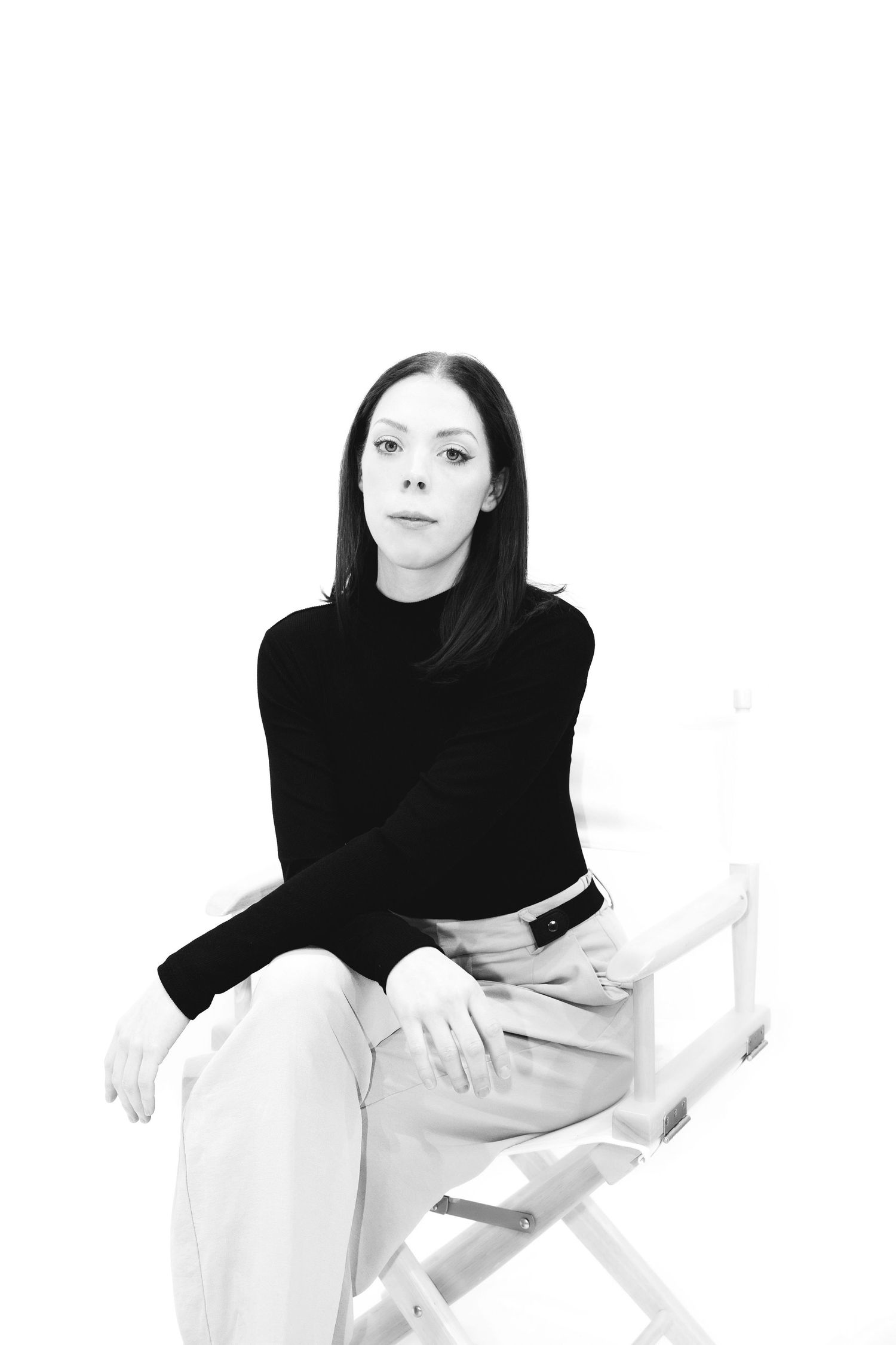 Minimalist black and white portrait of a woman seated confidently in a chair, dressed in a black long-sleeve top and light pants.