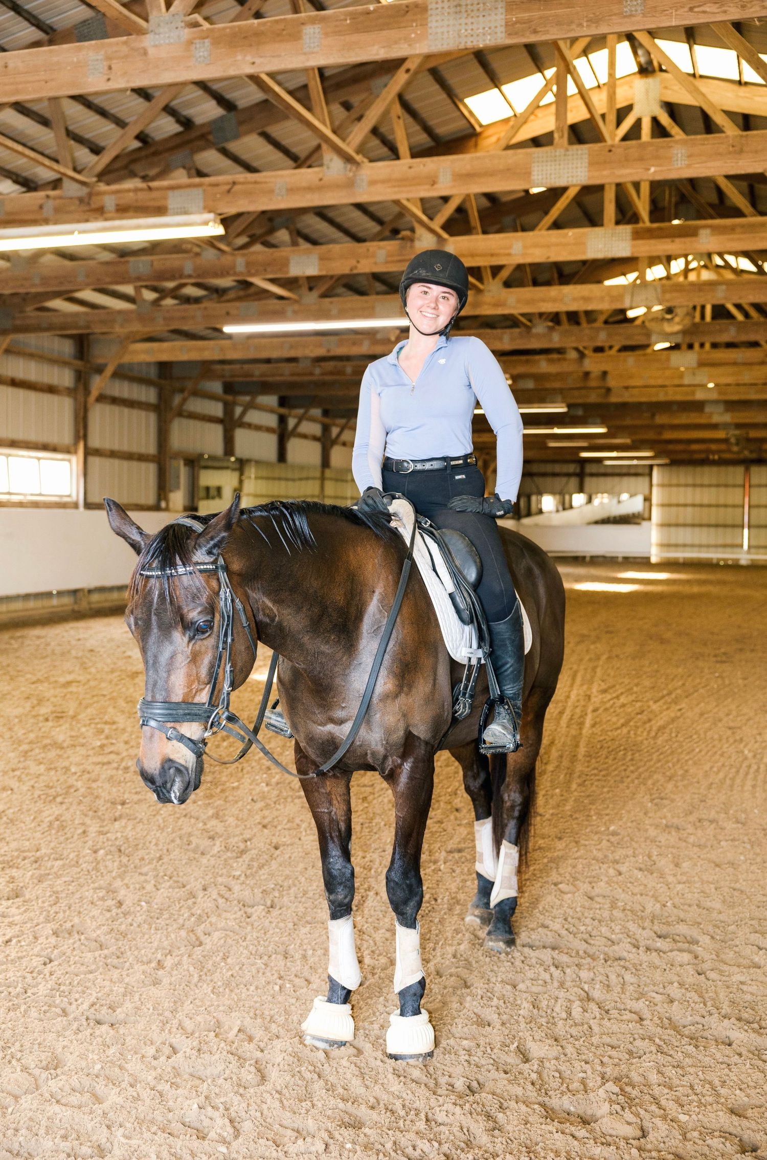Equestrian rider wearing a helmet sits on a brown horse inside an indoor riding arena with wooden beams and sandy footing.