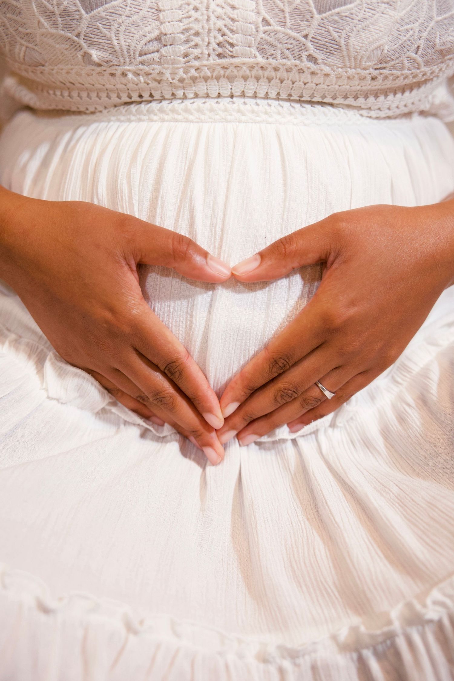 Close-up maternity portrait of hands forming a heart over a pregnant belly, dressed in a soft white lace gown.