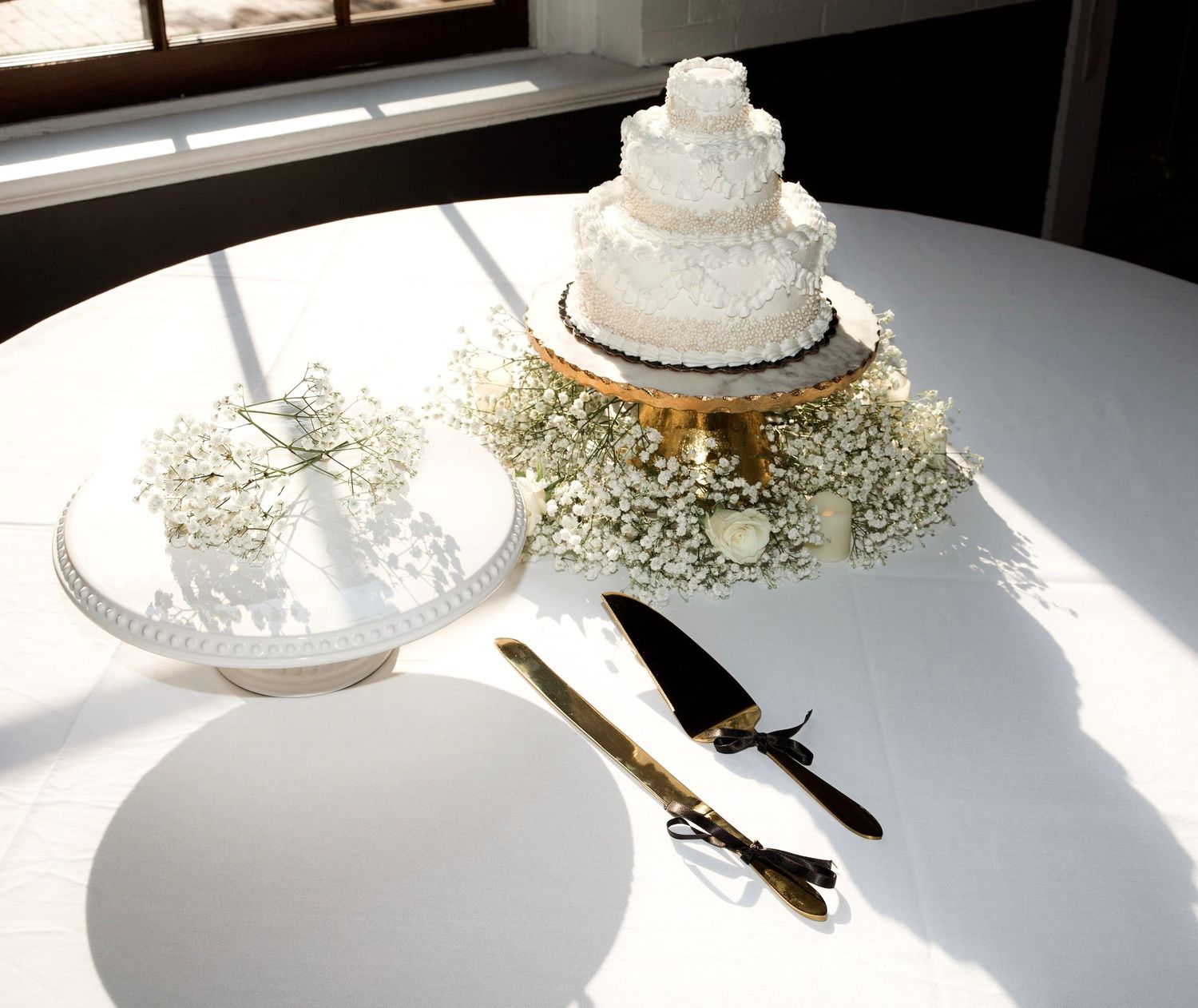 Three-tier white wedding cake styled with delicate floral details, cake stand, and gold serving utensils on a linen-covered table.
