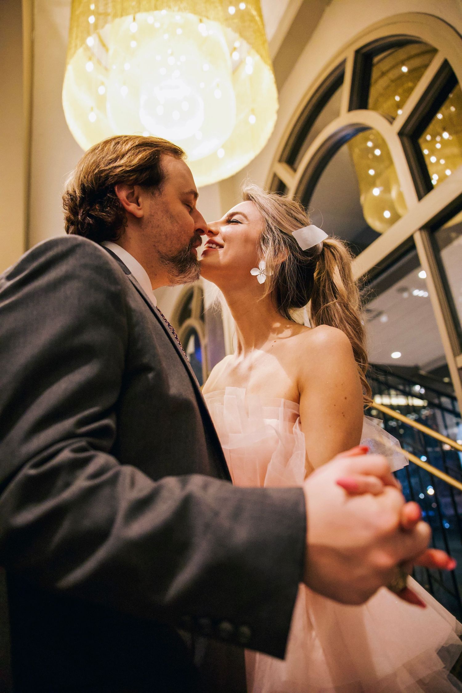 Couple share a quiet kiss while holding hands indoors, framed by warm light and soft architectural details.