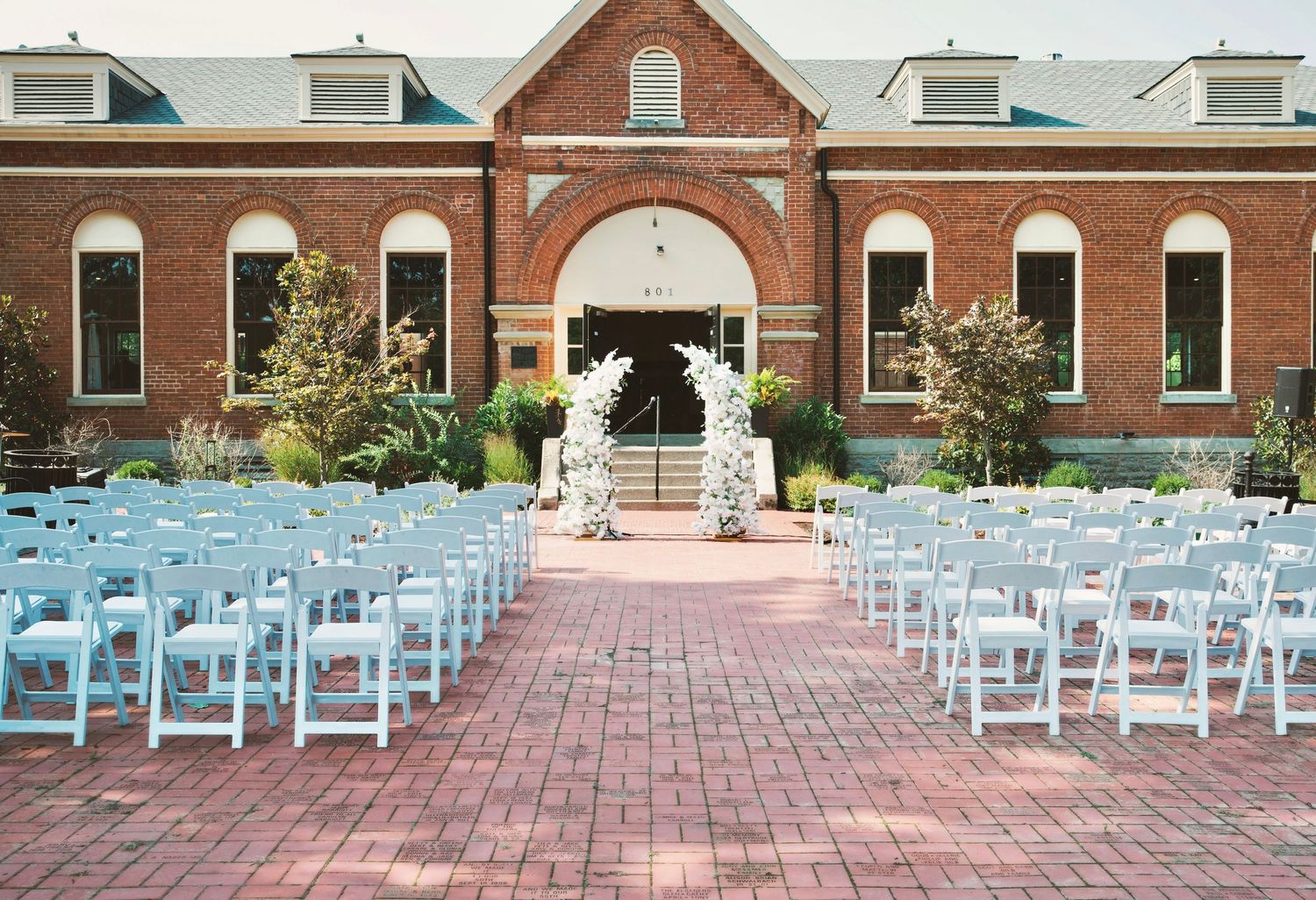 Outdoor wedding ceremony setup with white chairs facing a floral arch in front of a red brick building on a sunny day.