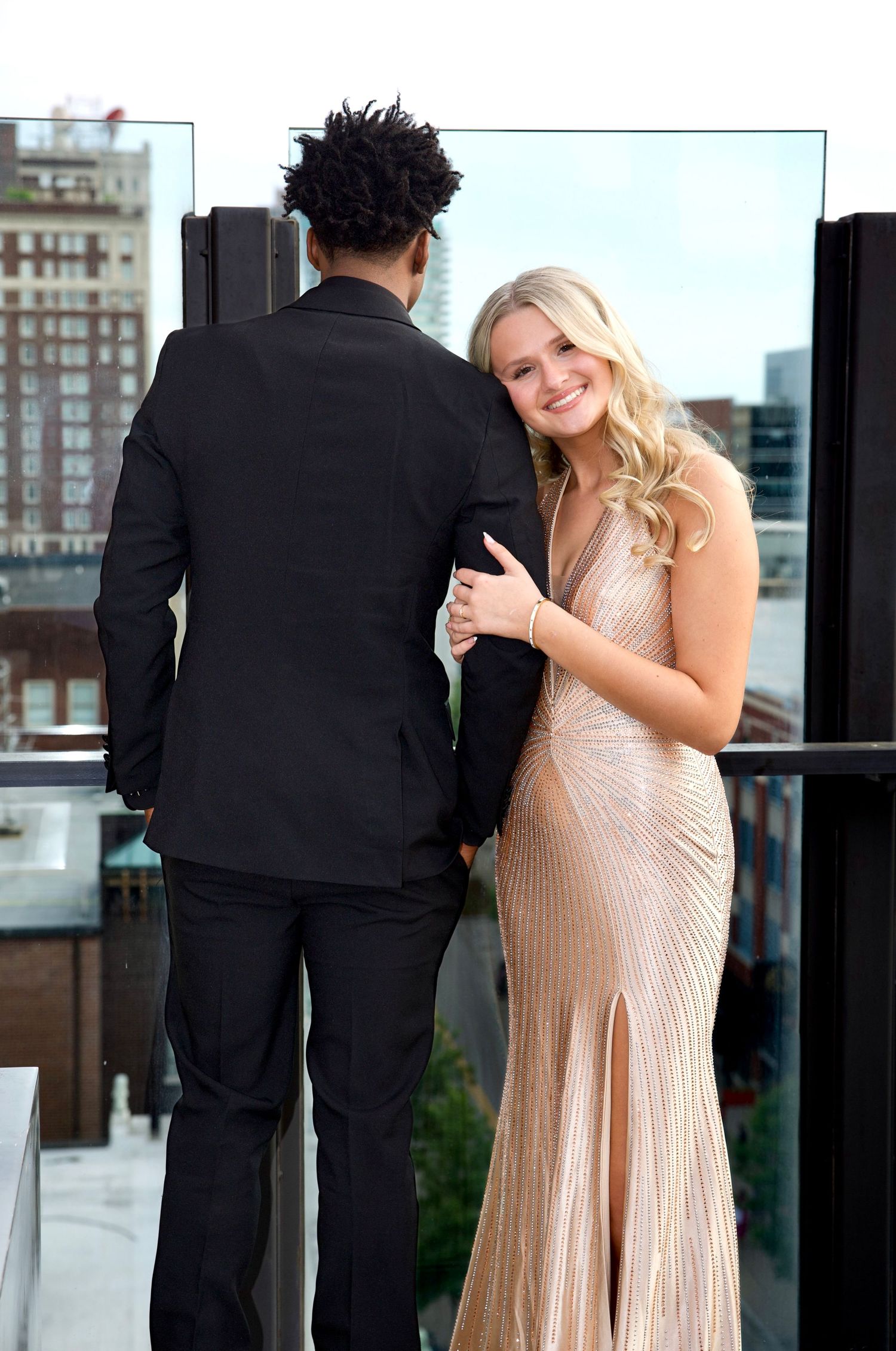 Formally dressed couple stands on a rooftop balcony, with city buildings in the background during an elegant portrait session.