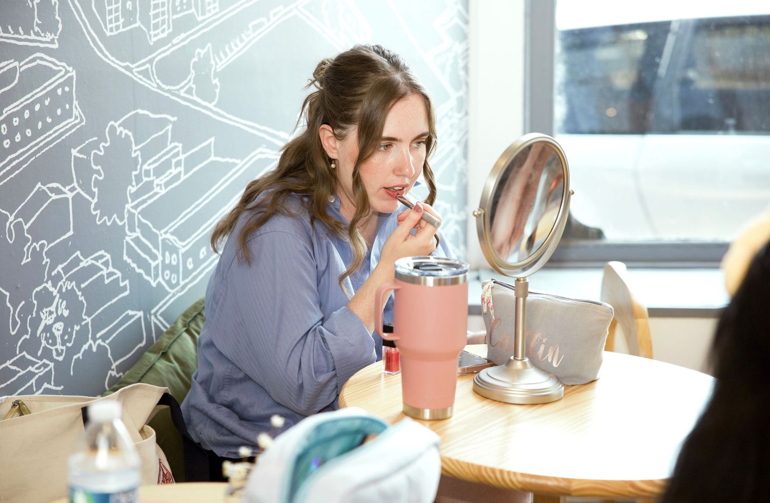Woman applies lipstick while seated at a table, using a mirror in a bright, modern indoor setting during event preparation.