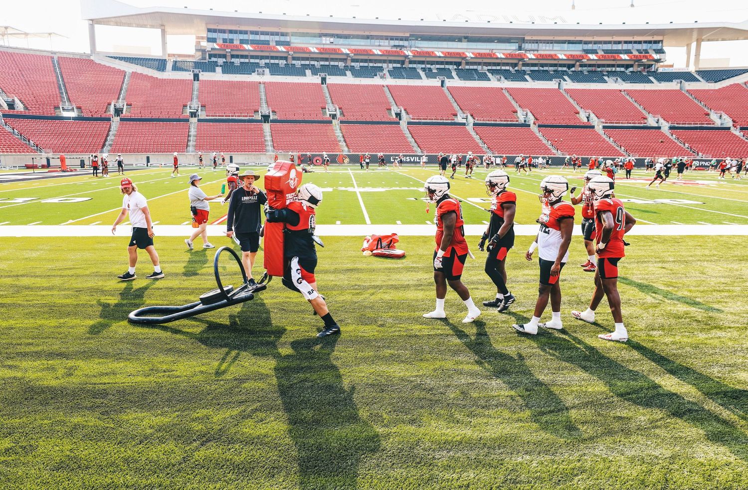 Athletes in football helmets and pads line up for a blocking drill during practice on a sunny stadium field.