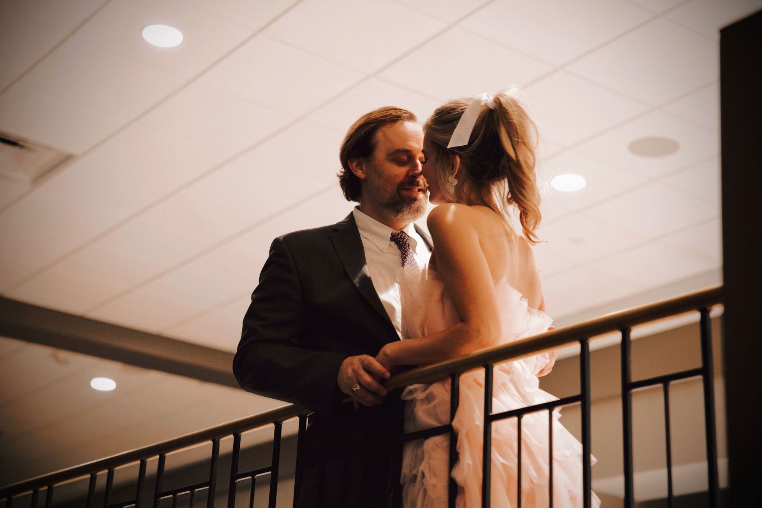 Couple share a quiet, intimate moment while leaning together on a balcony, captured in warm indoor light.