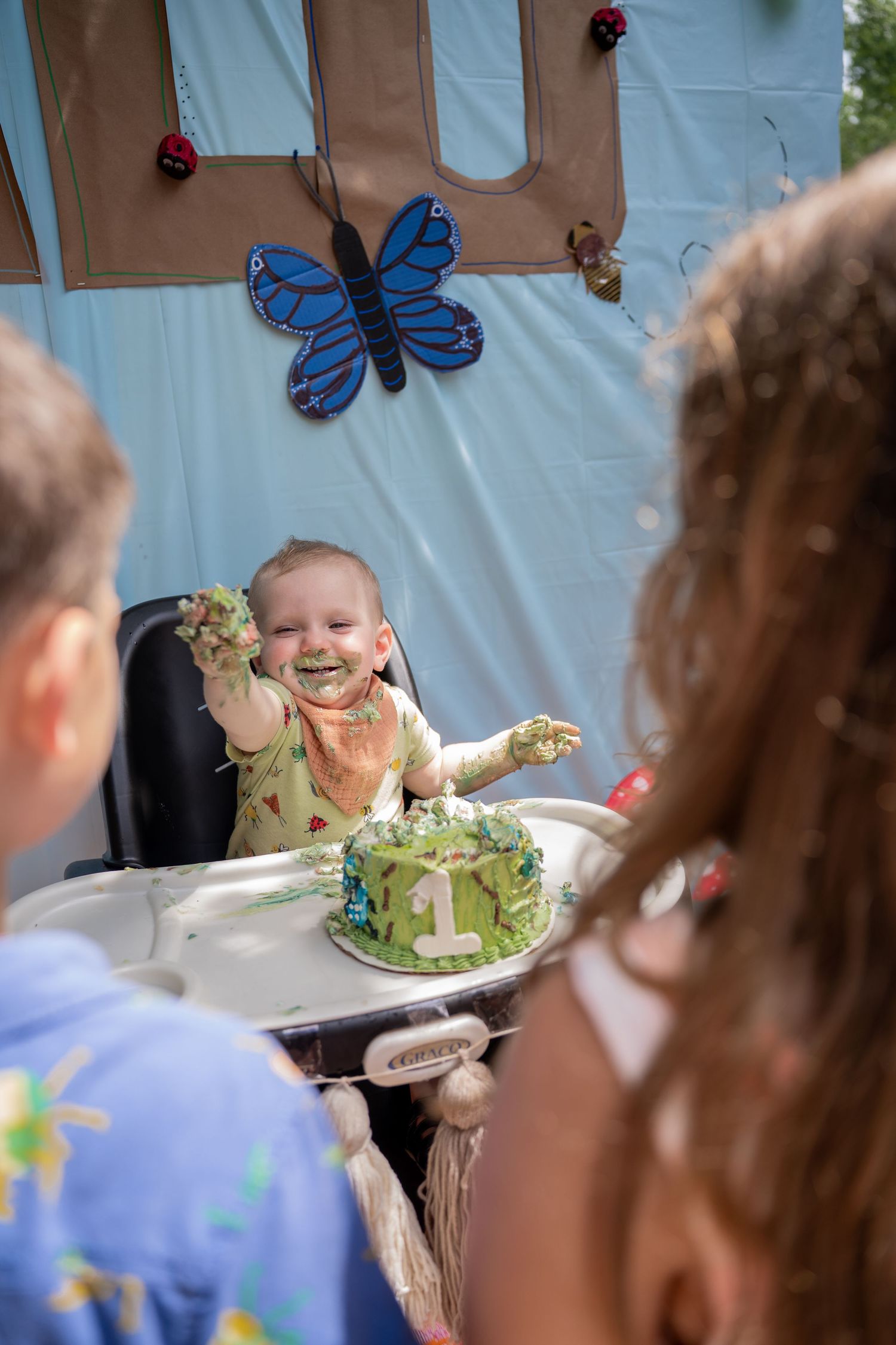 Toddler holding up cake to two children watching