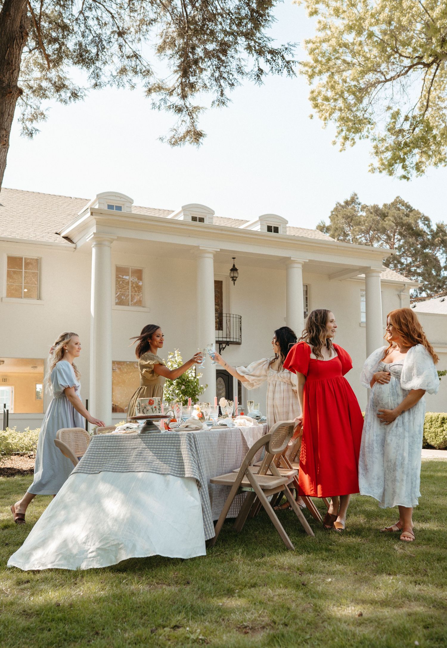 Women in colorful dresses gather around an outdoor table at a garden baby shower party.