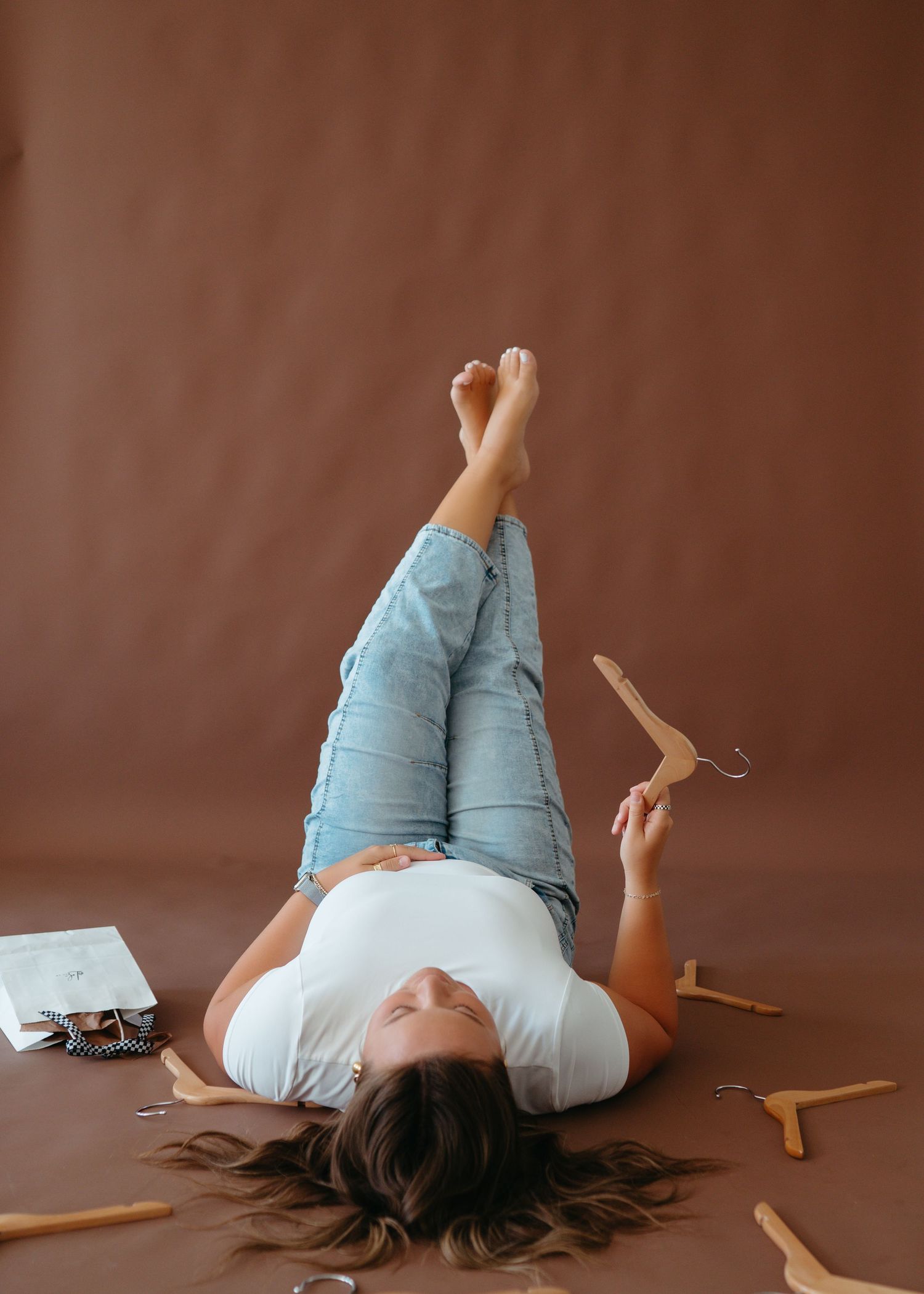 A woman in jeans lies on a brown studio floor surrounded by wooden hangers.