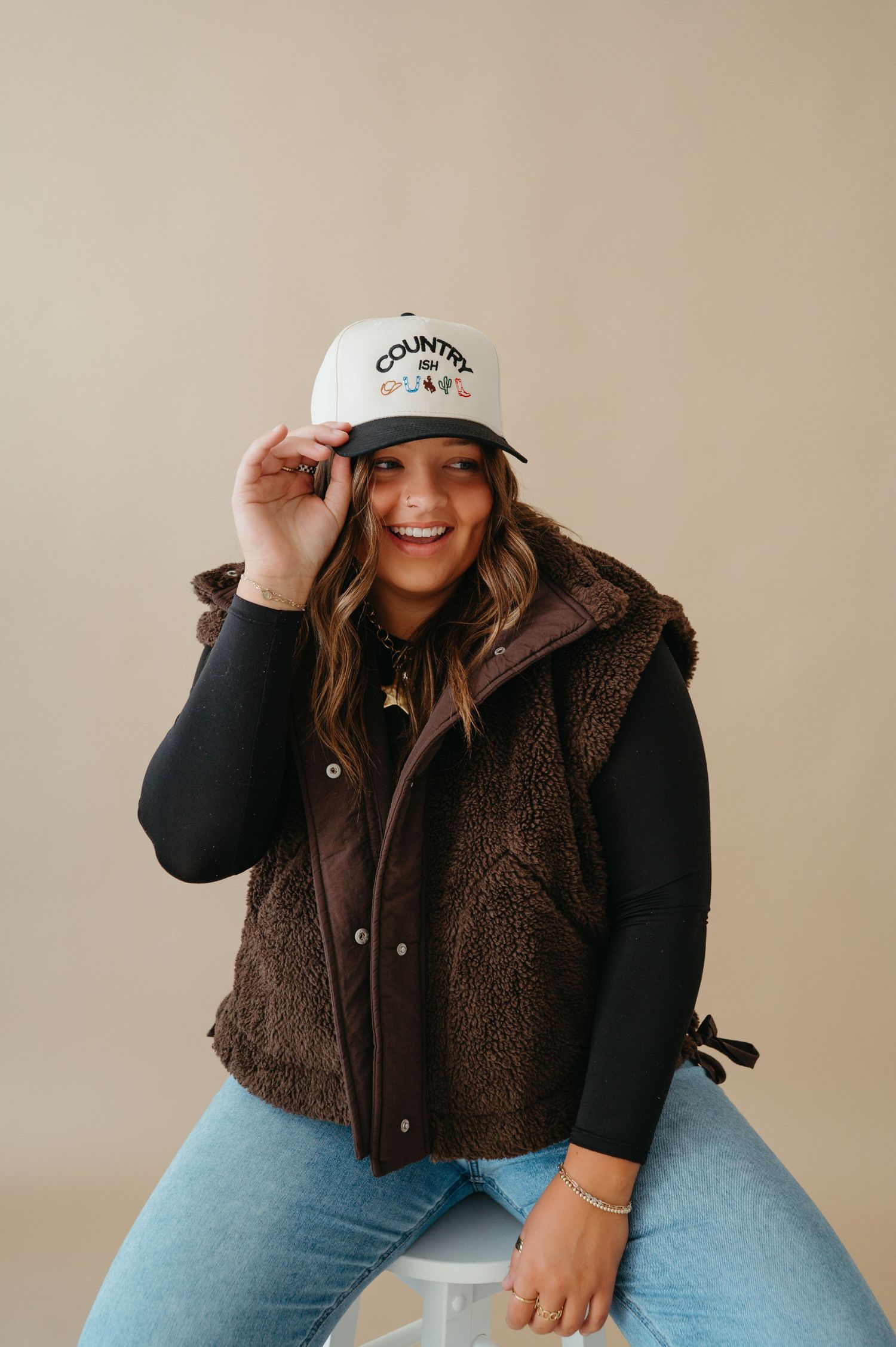 Cheerful woman in brown sherpa vest adjusting a Country Soul trucker hat while sitting on a white stool.