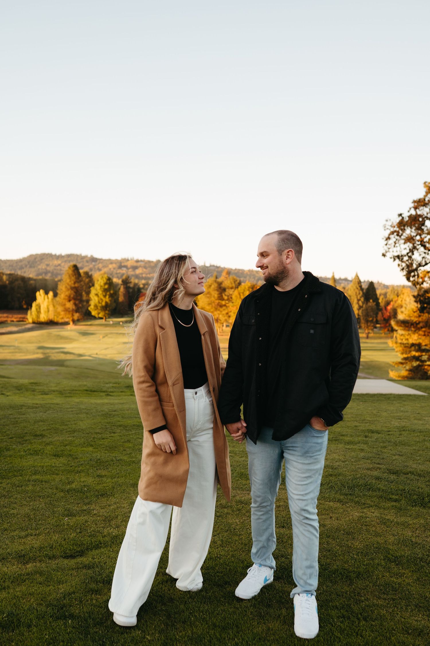 Happy couple holding hands and smiling at each other on a green golf course during golden hour autumn sunset.