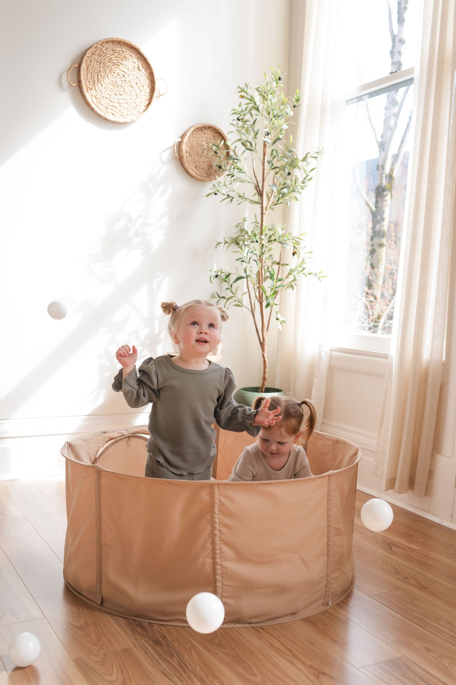 Two young children play with white balls inside a tan fabric ball pit indoors.