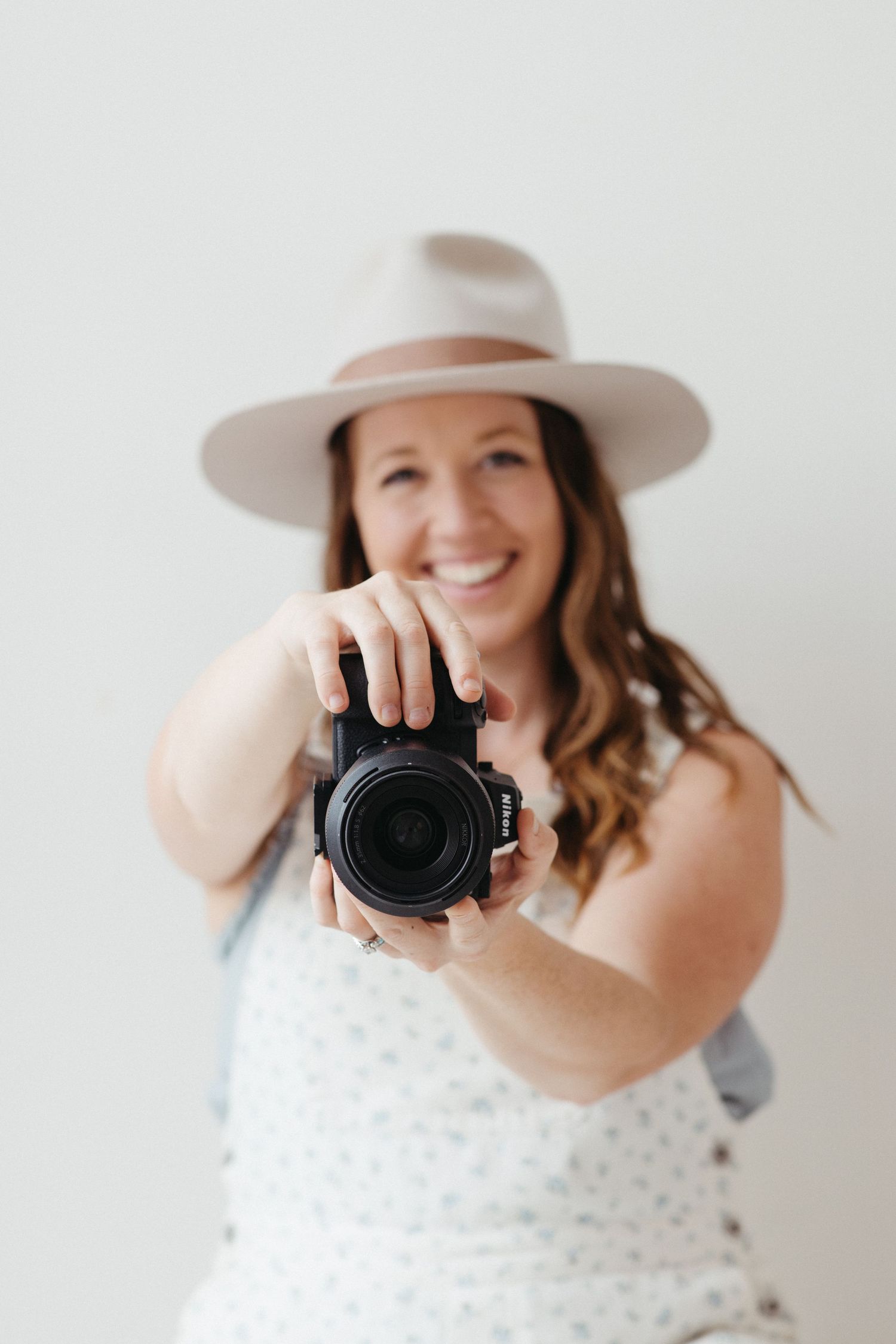 Smiling woman in a wide-brim hat holding a Nikon camera toward the lens against a white background.