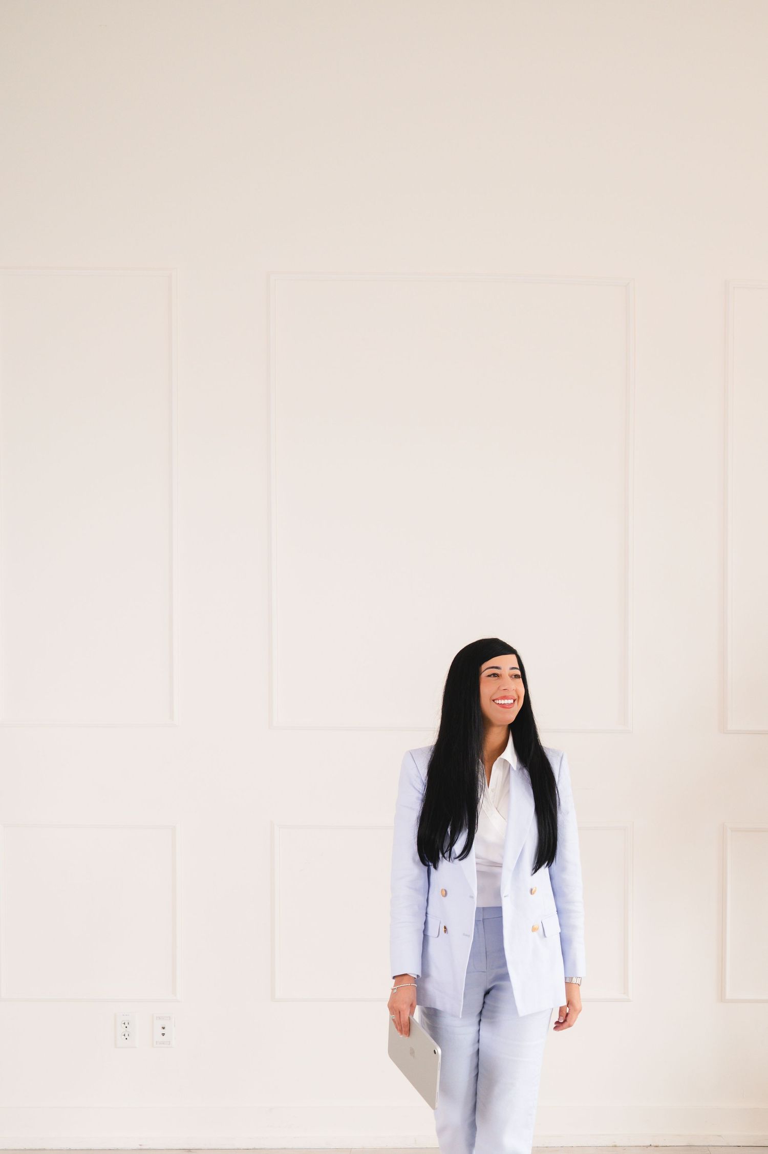 Professional woman in light blue suit smiling while standing against white paneled wall.