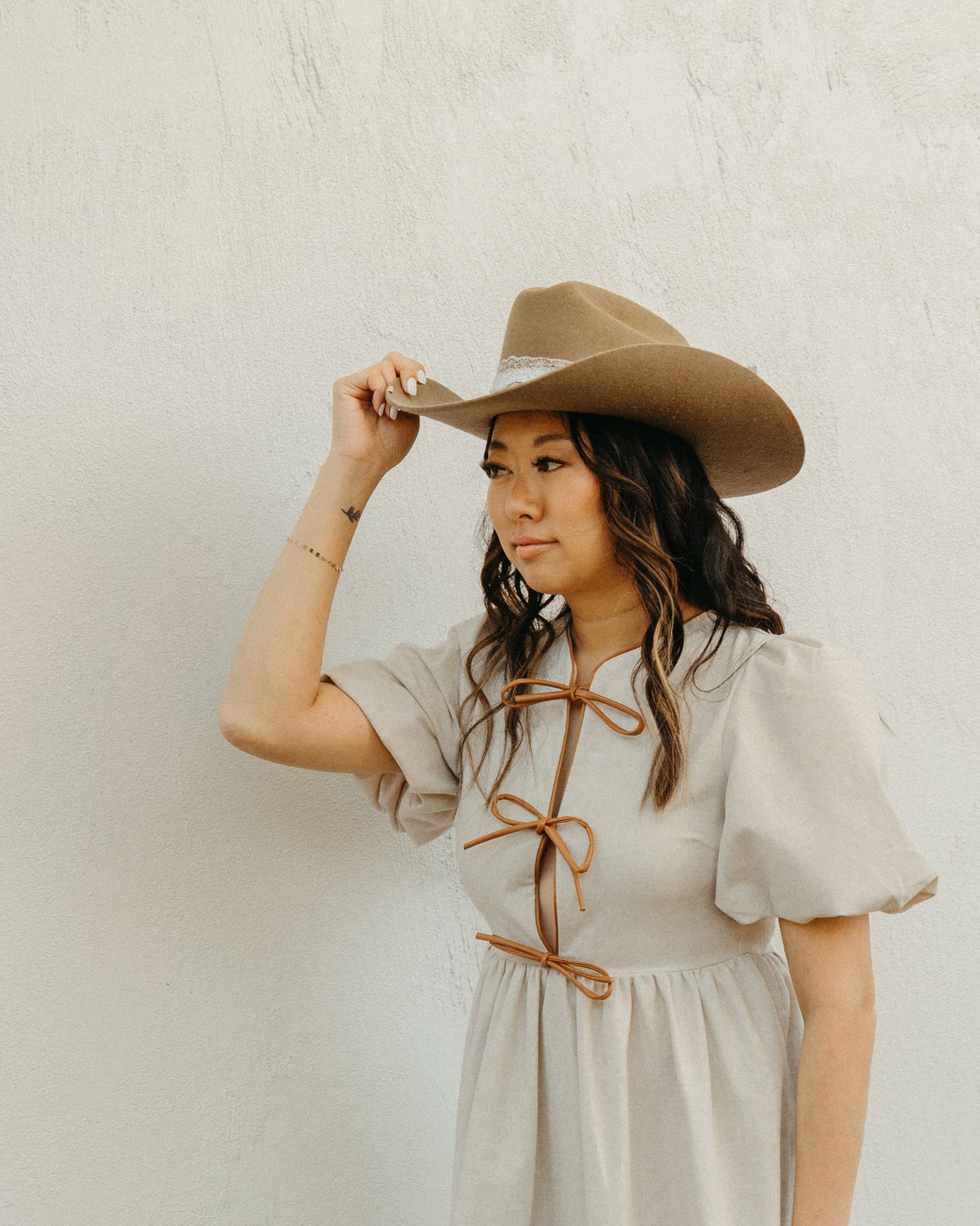 Stylish woman in a tan wide-brim cowboy hat and linen dress poses against a white wall.