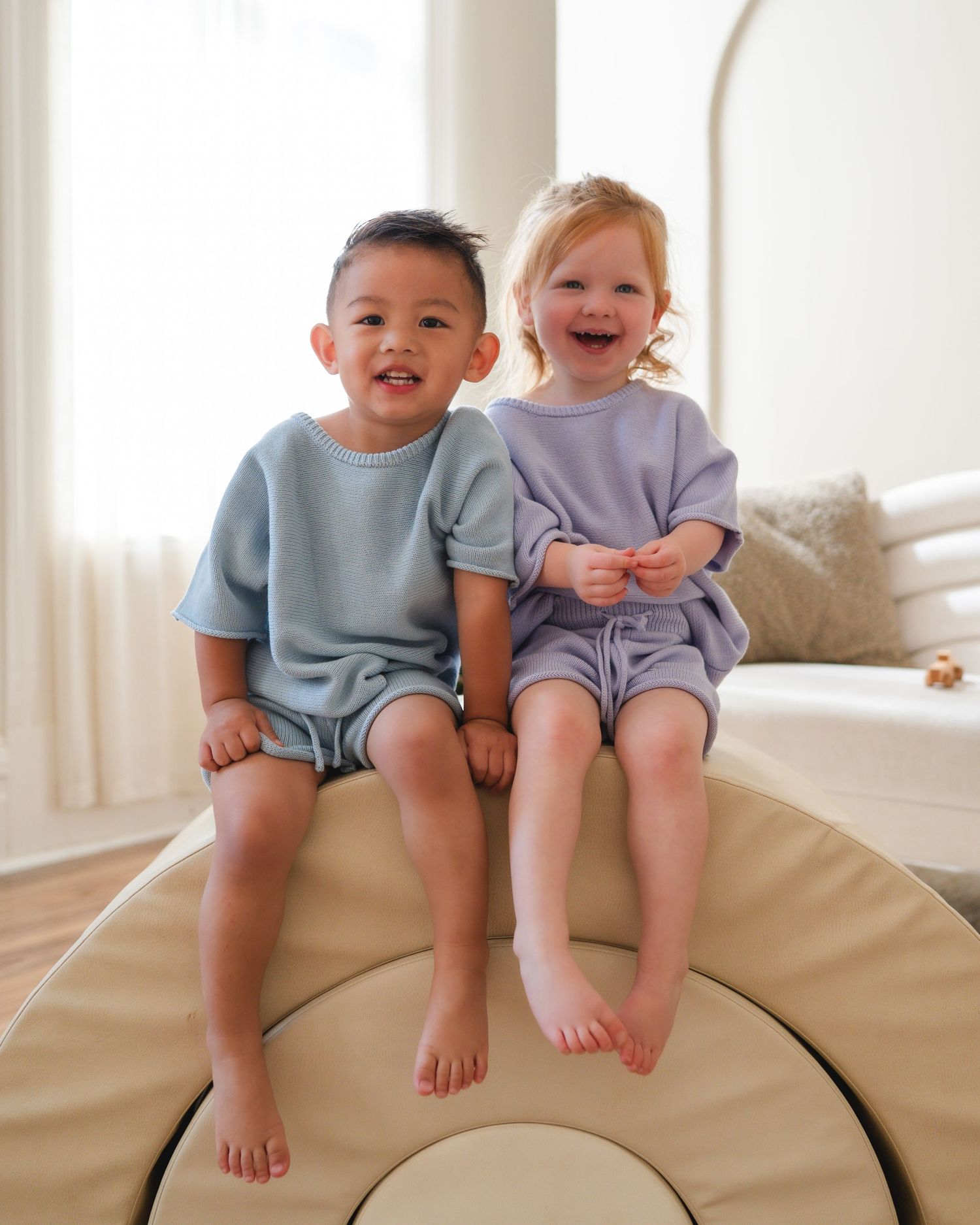 Two smiling toddlers in blue and lavender waffle knit sets sit together on a cushion.