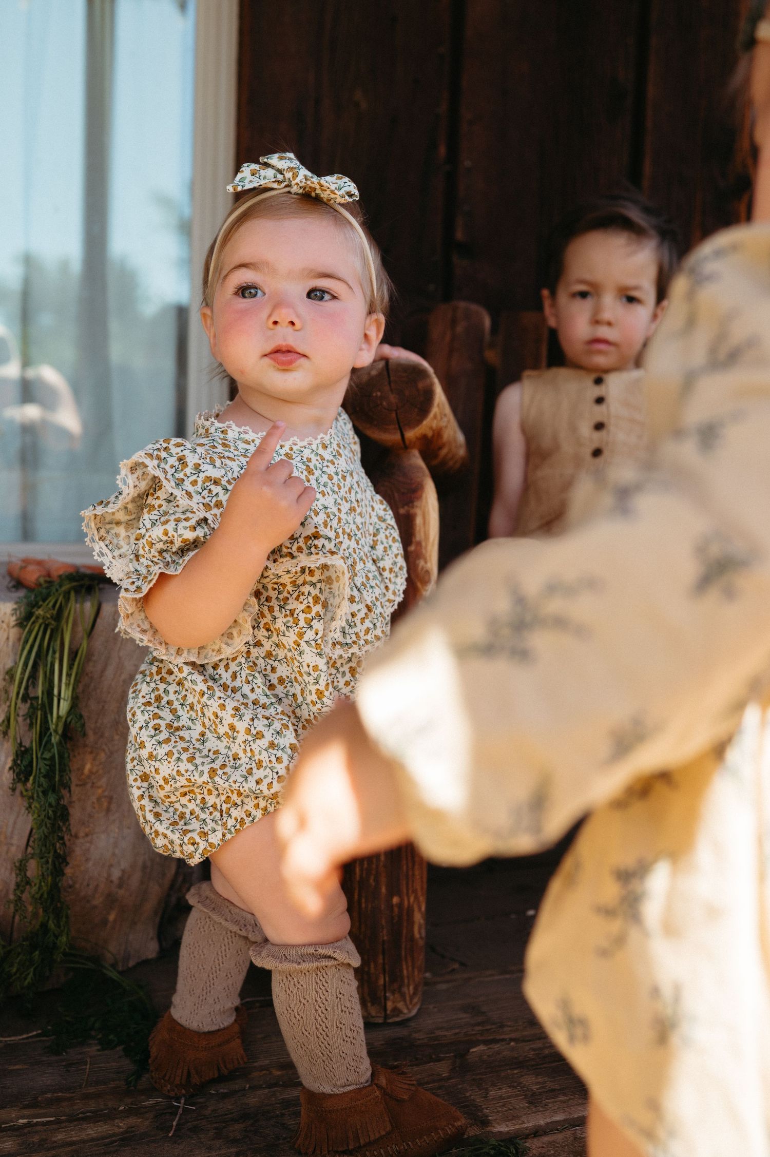 Adorable baby girl in floral romper and bow headband stands near a rustic wooden rocking horse.