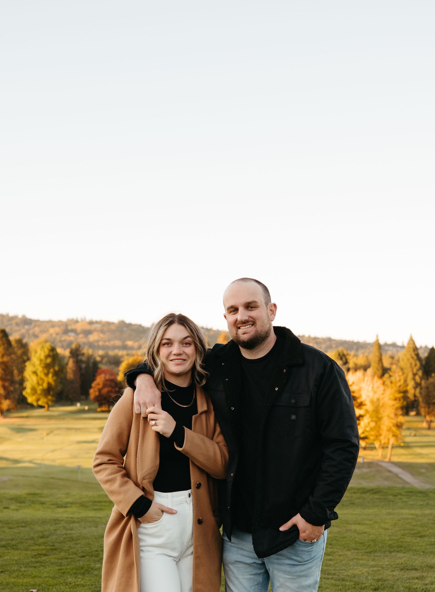 Smiling couple posing together on a lush green golf course with vibrant autumn foliage in the background.