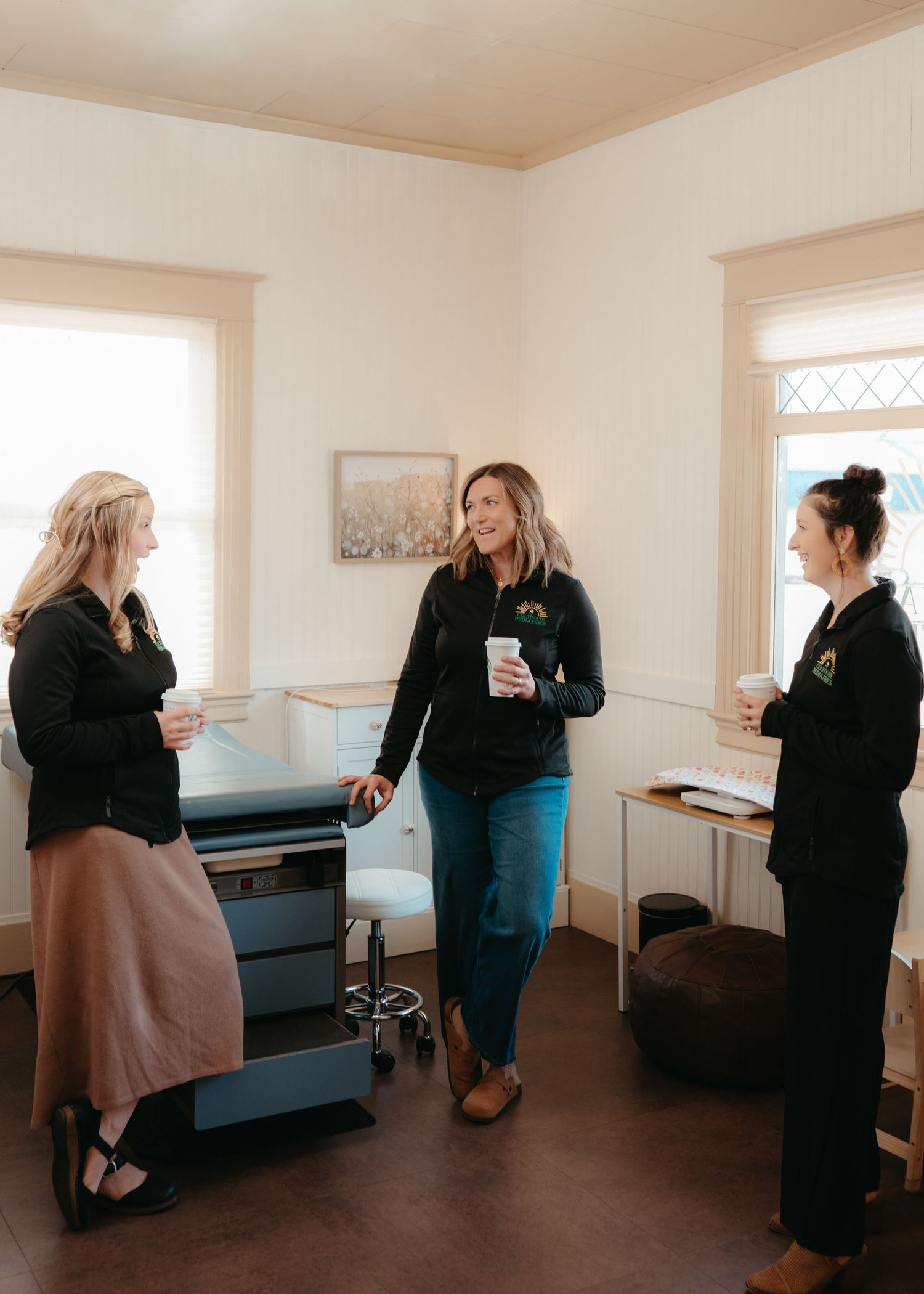 Three women in matching black jackets chatting and holding coffee cups indoors.