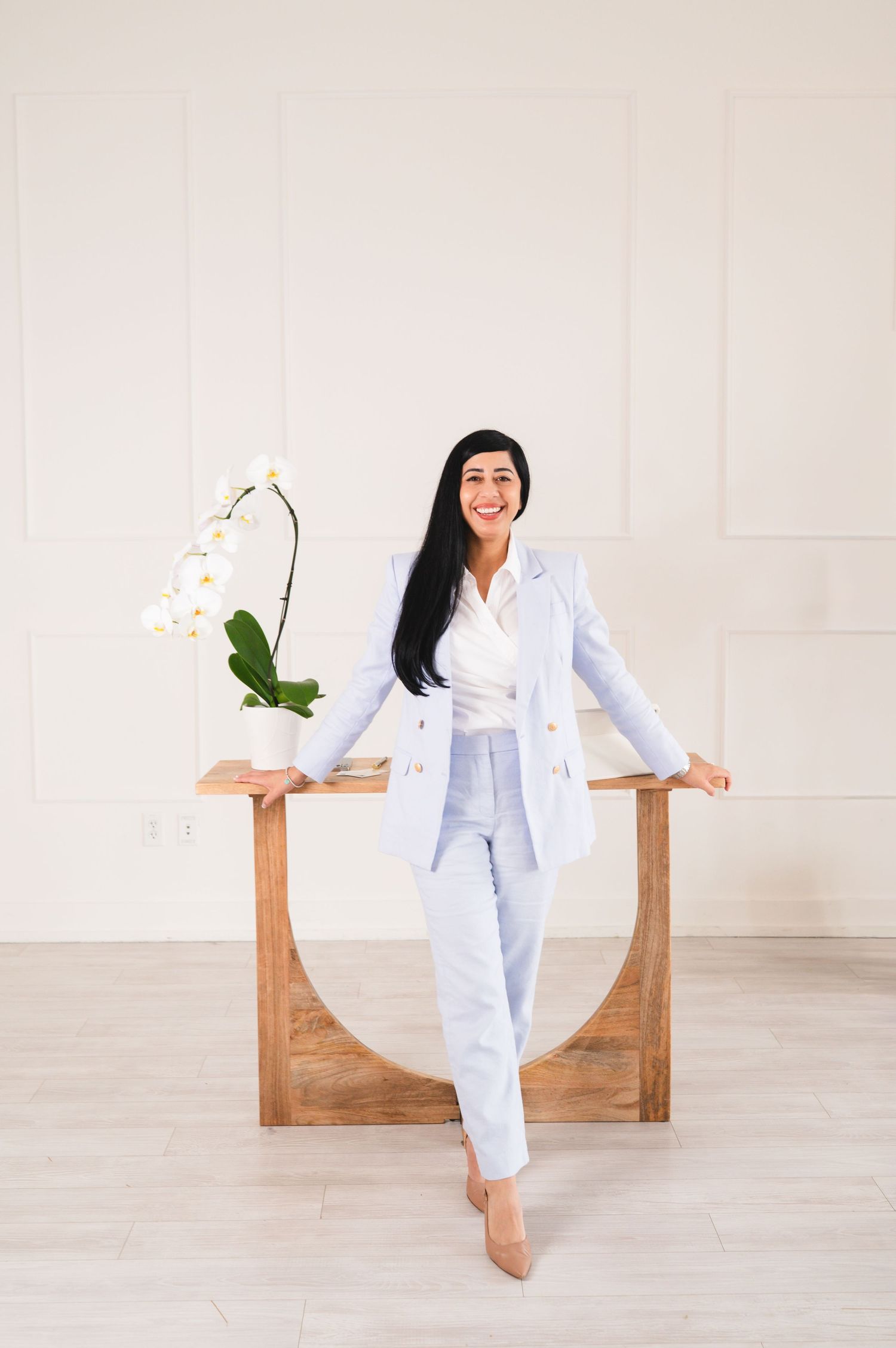 Professional woman in light blue suit posing confidently beside a wooden table with white orchids in a bright studio.