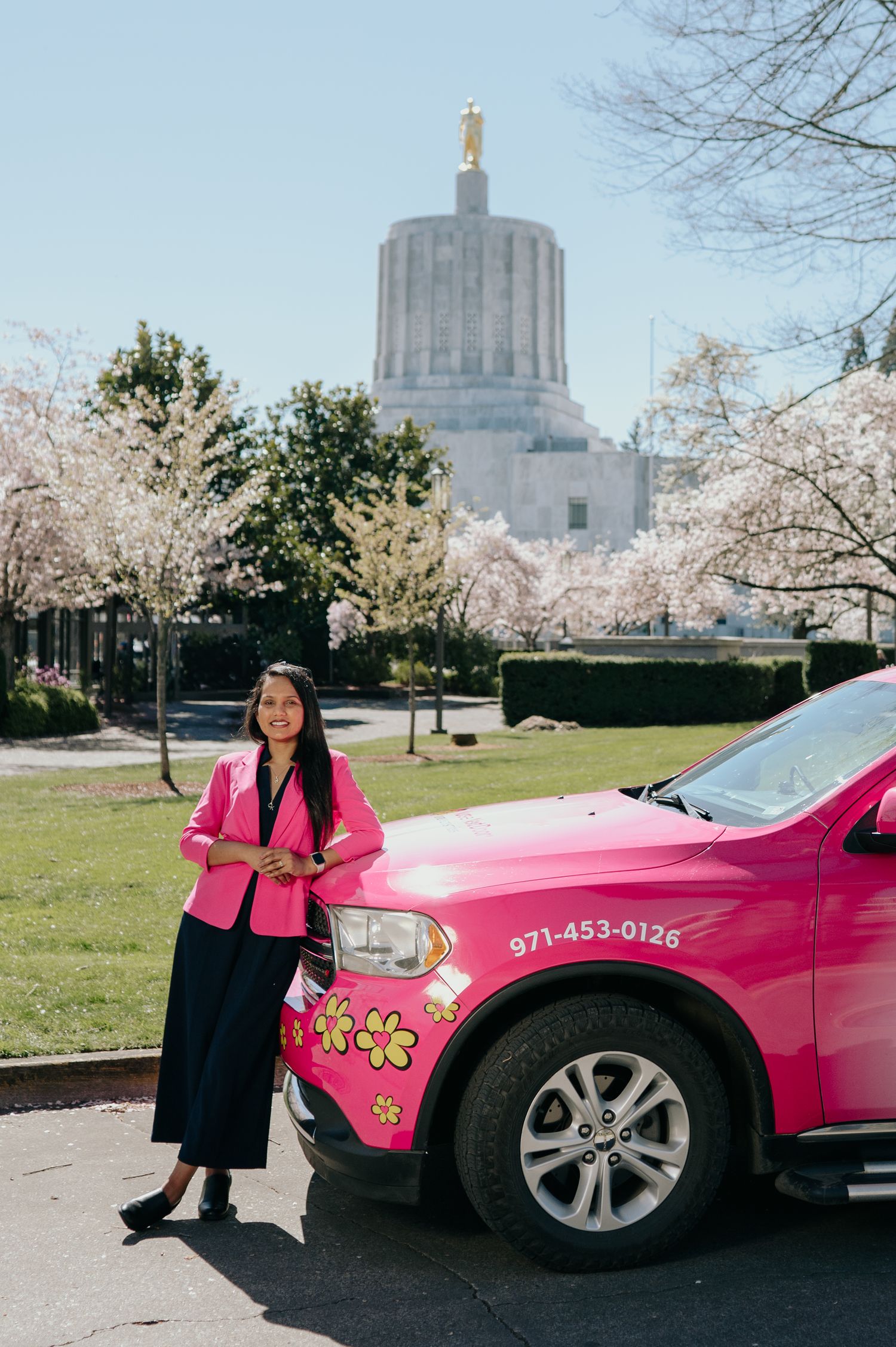 Woman in pink blazer standing beside bright pink floral car near Oregon State Capitol building.
