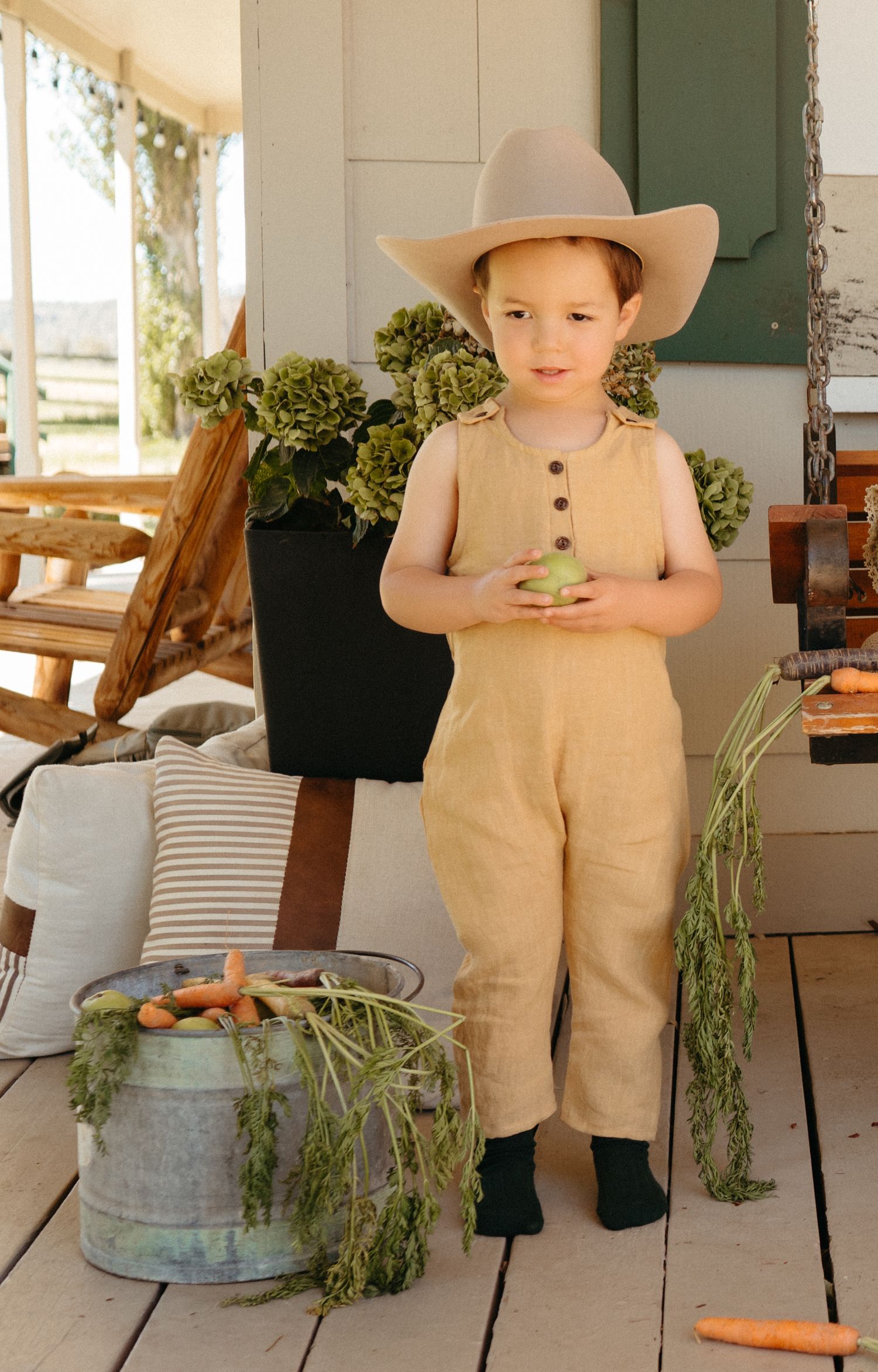 A young child in a tan linen romper and wide-brim hat stands on a rustic porch.