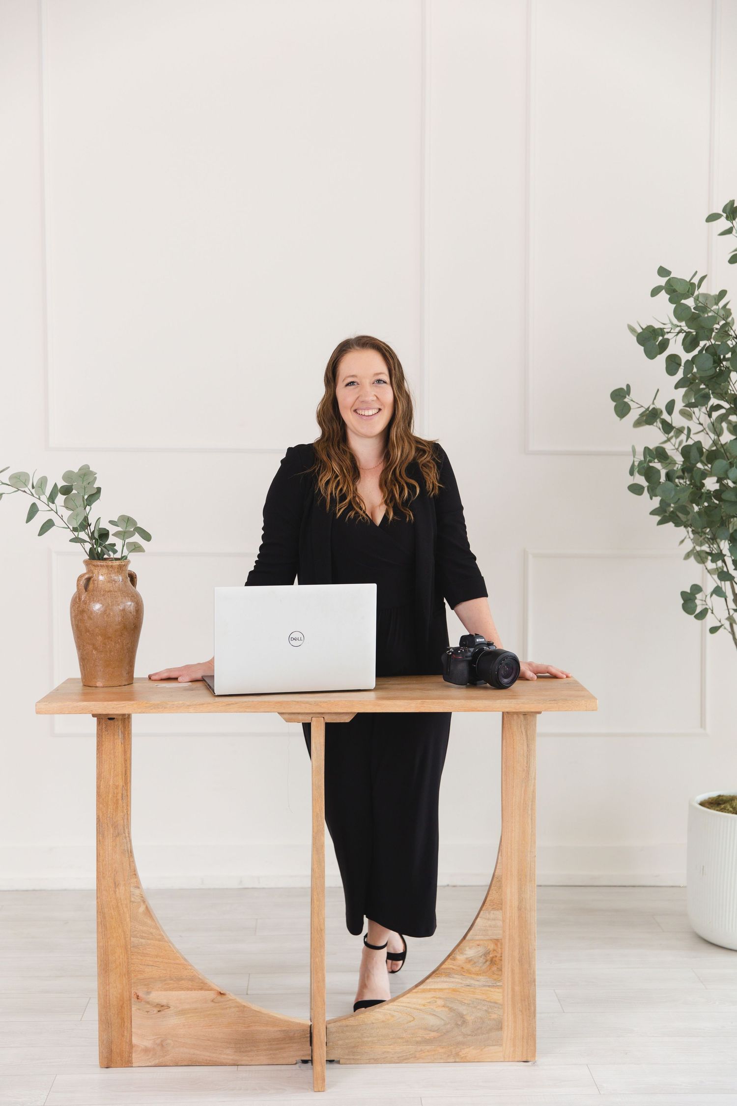 Woman in black dress smiling at wooden desk with Dell laptop and camera in bright white studio.