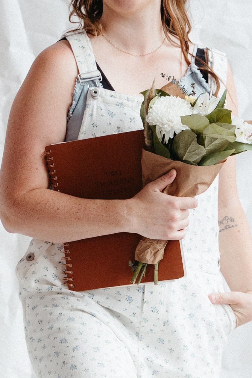 Woman in floral overalls holding brown spiral notebook and white flower bouquet.