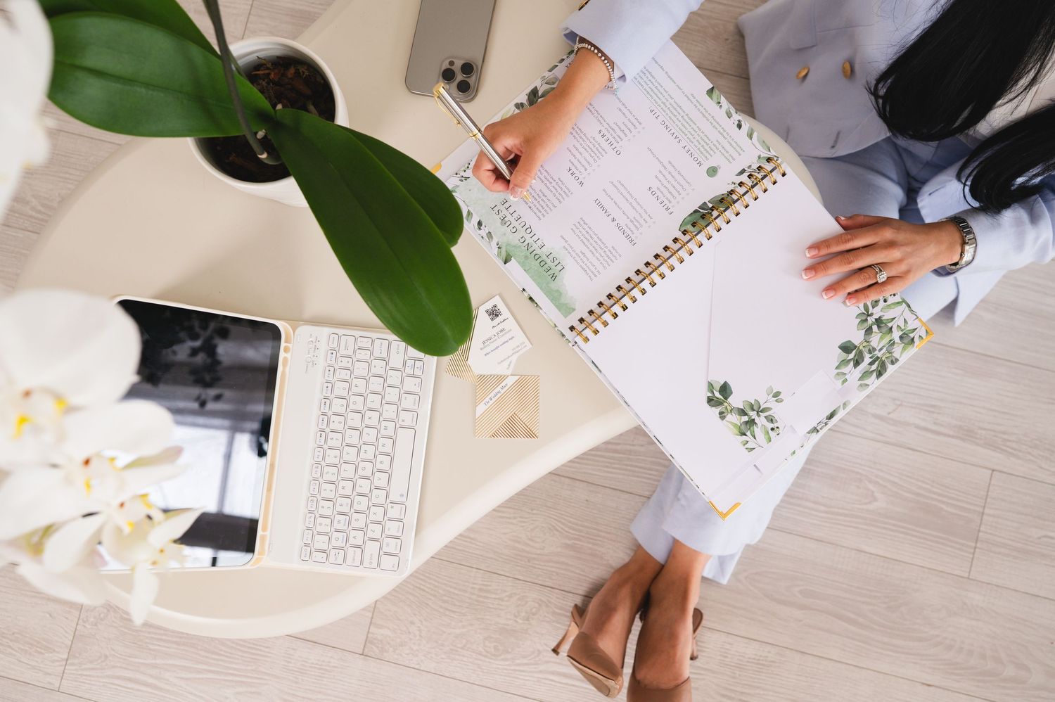 Overhead view of a woman writing in a spiral planner at a white desk with a laptop, orchid plant, and white flowers.