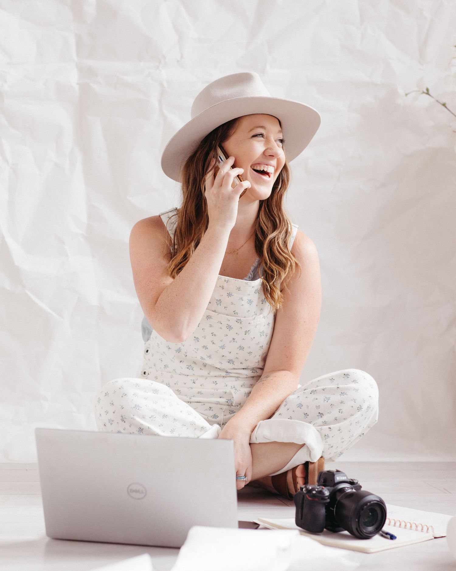 A cheerful woman in a floral jumpsuit and hat laughs while on her smartphone.