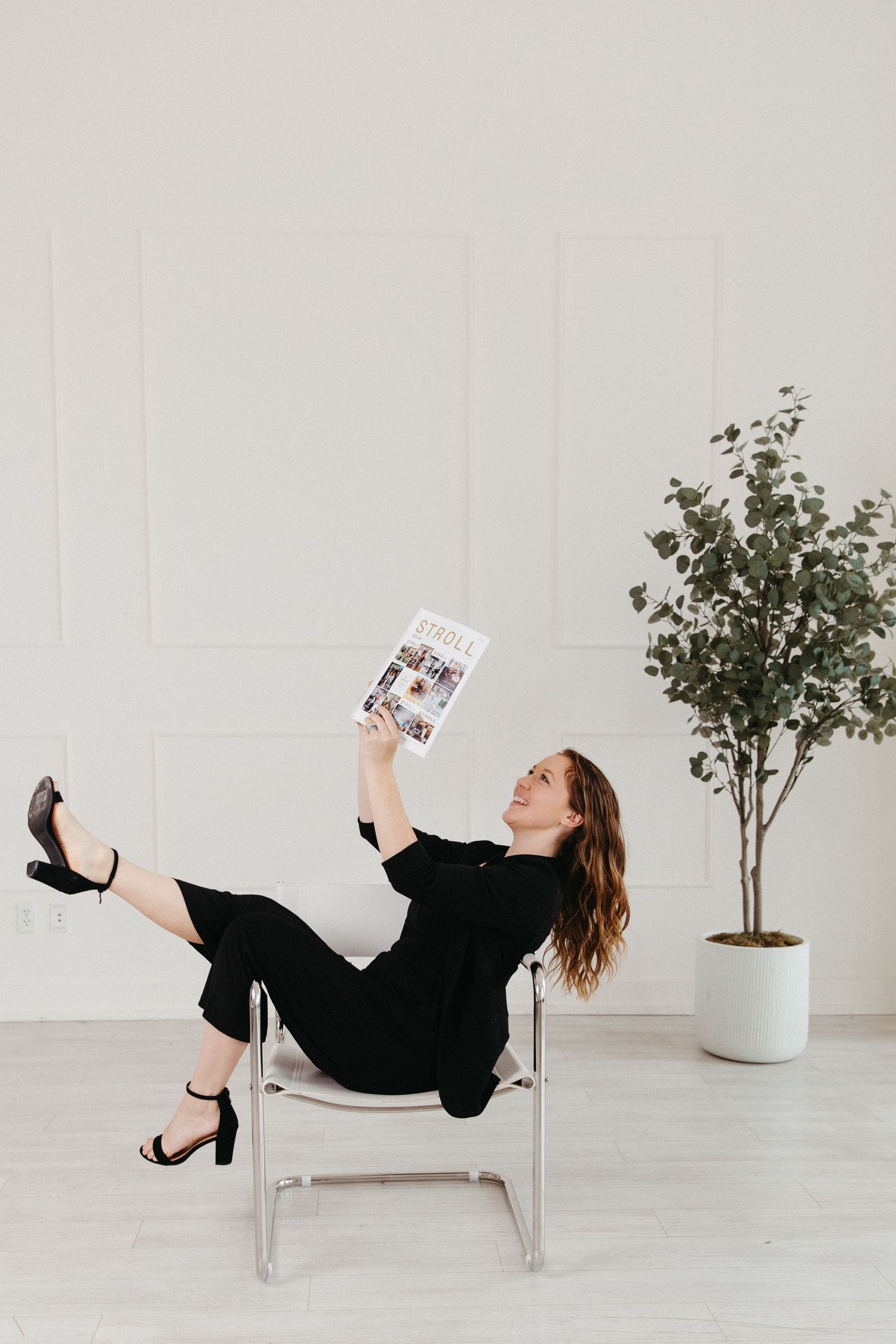 Smiling woman in black jumpsuit lounging in chair holding Stroll magazine in bright white room.
