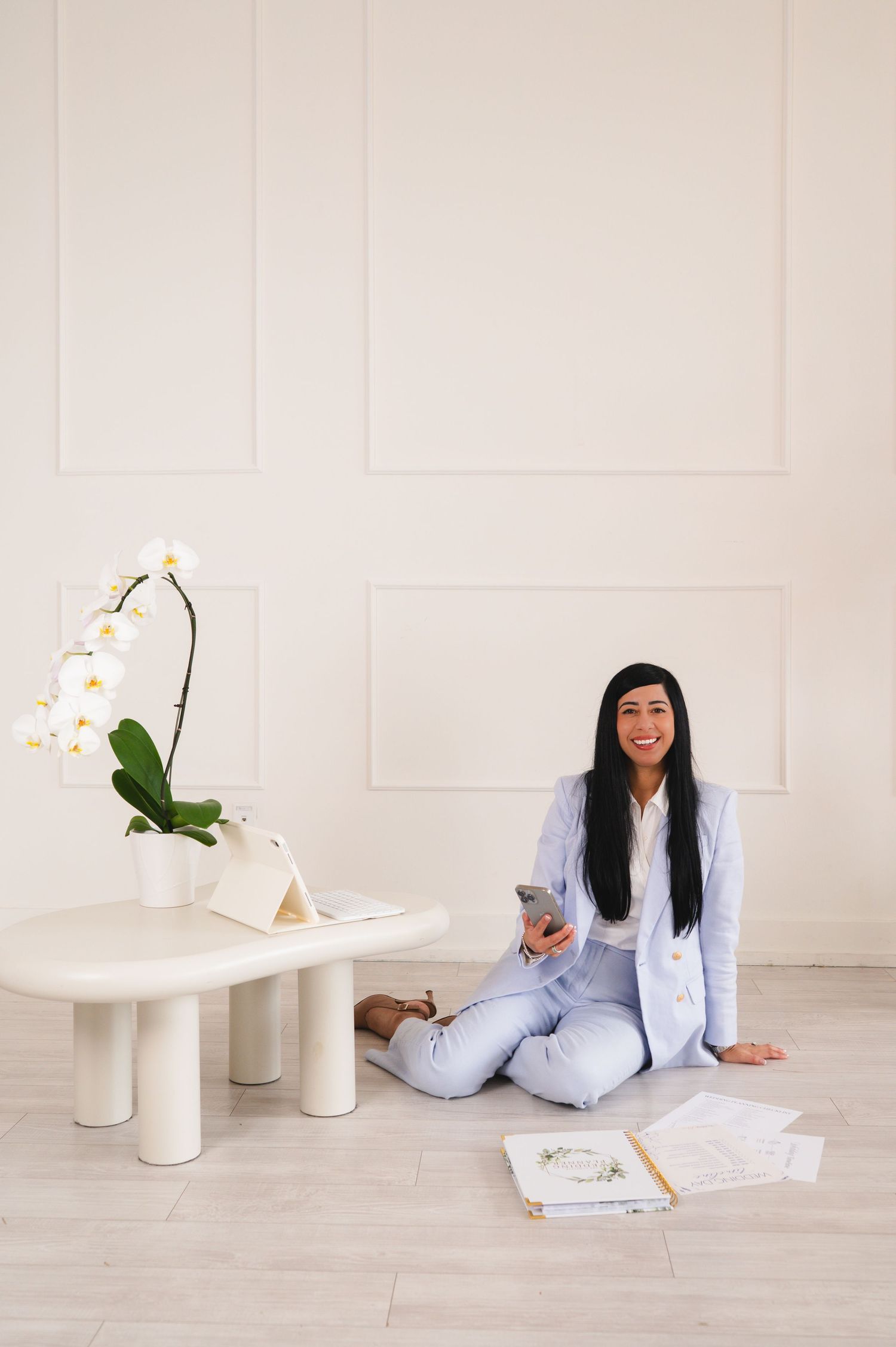 Woman in blue suit sitting on floor with phone surrounded by papers near white orchid.