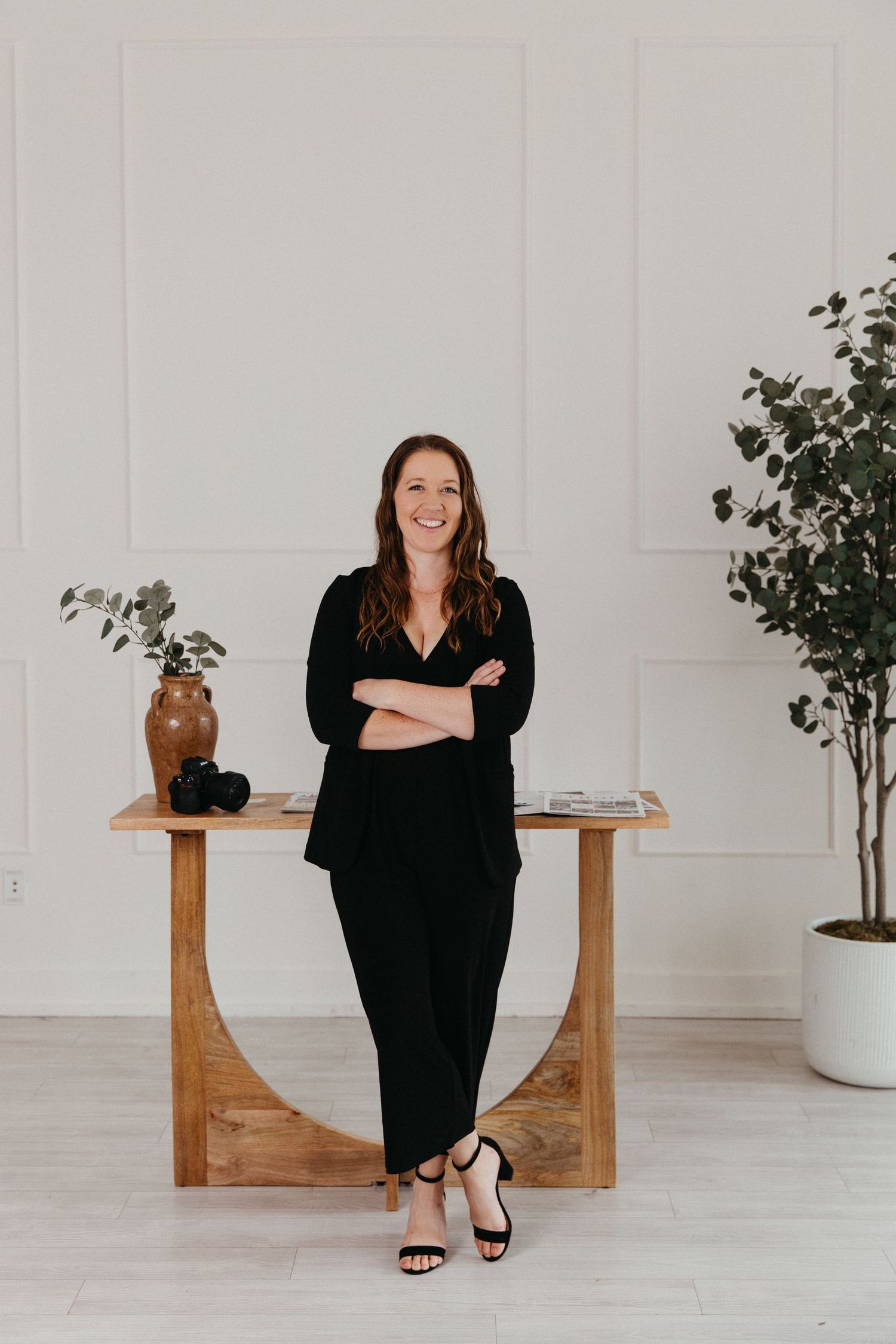 Professional woman in black jumpsuit leaning against wooden table in elegant white studio space.