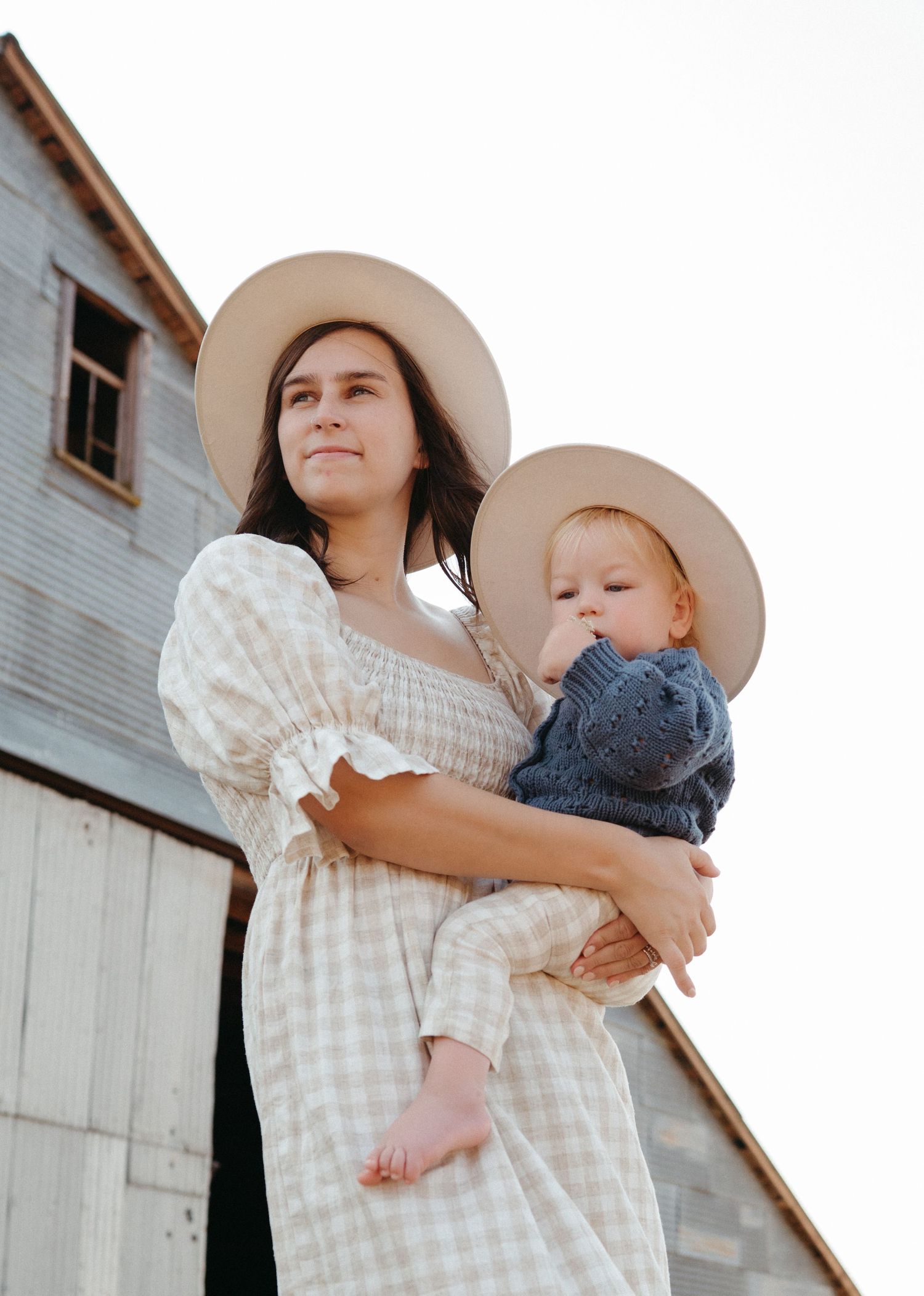 Mother and baby wearing matching tan hats pose together in front of a rustic barn.