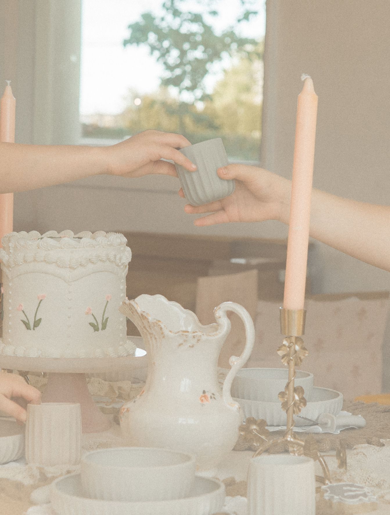 Hands exchanging a small ceramic cup over an elegant white cake and candle tablescape.