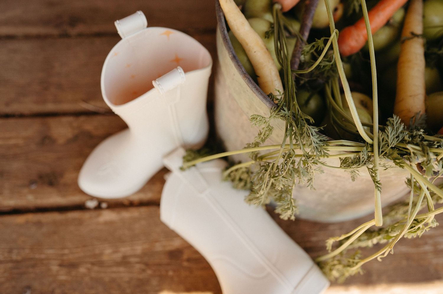 White ceramic cowboy boots used as vases holding fresh carrots and herbs on rustic wood.