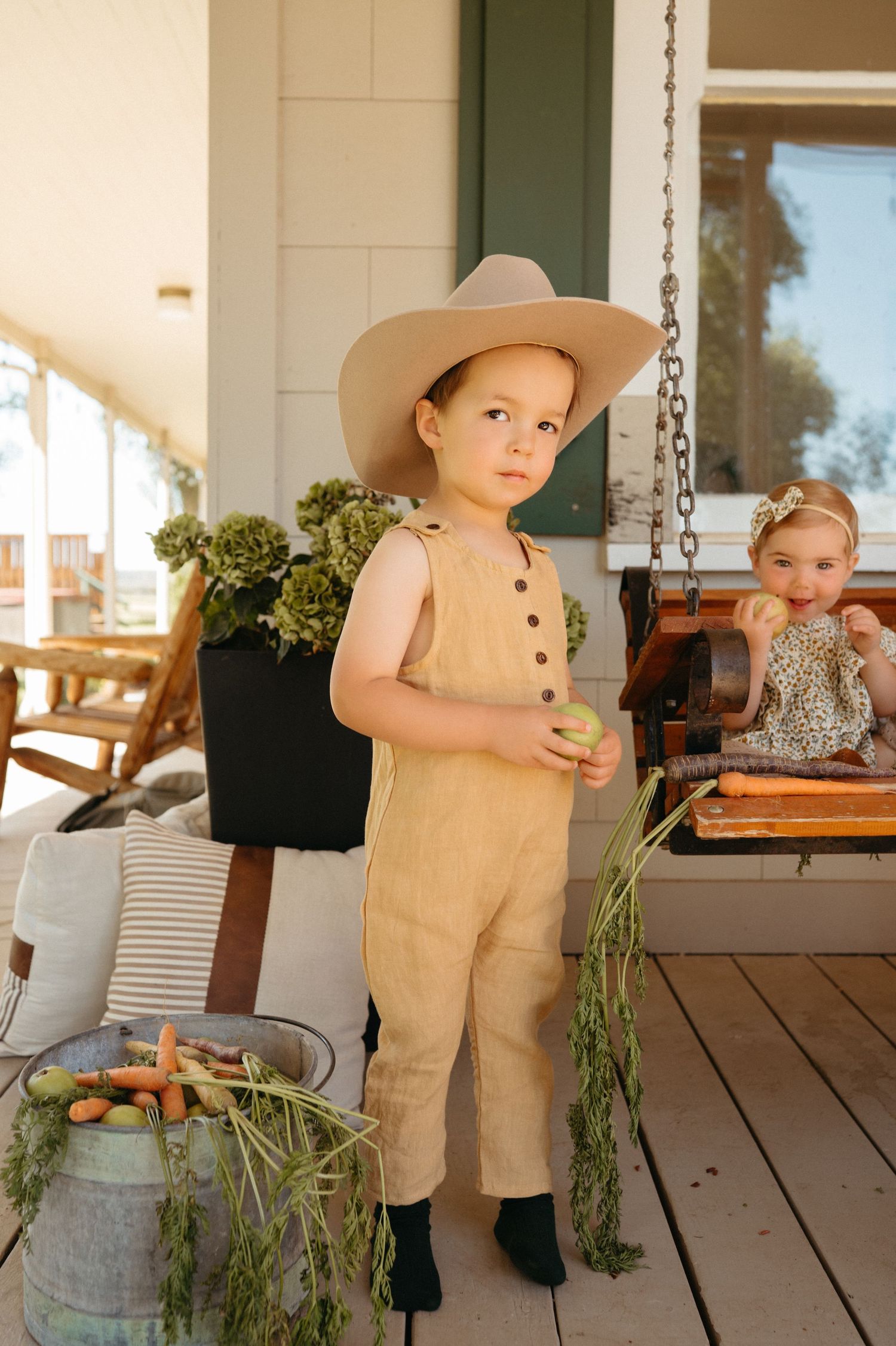 Toddler in tan overalls and cowboy hat holding apple on a farmhouse porch.